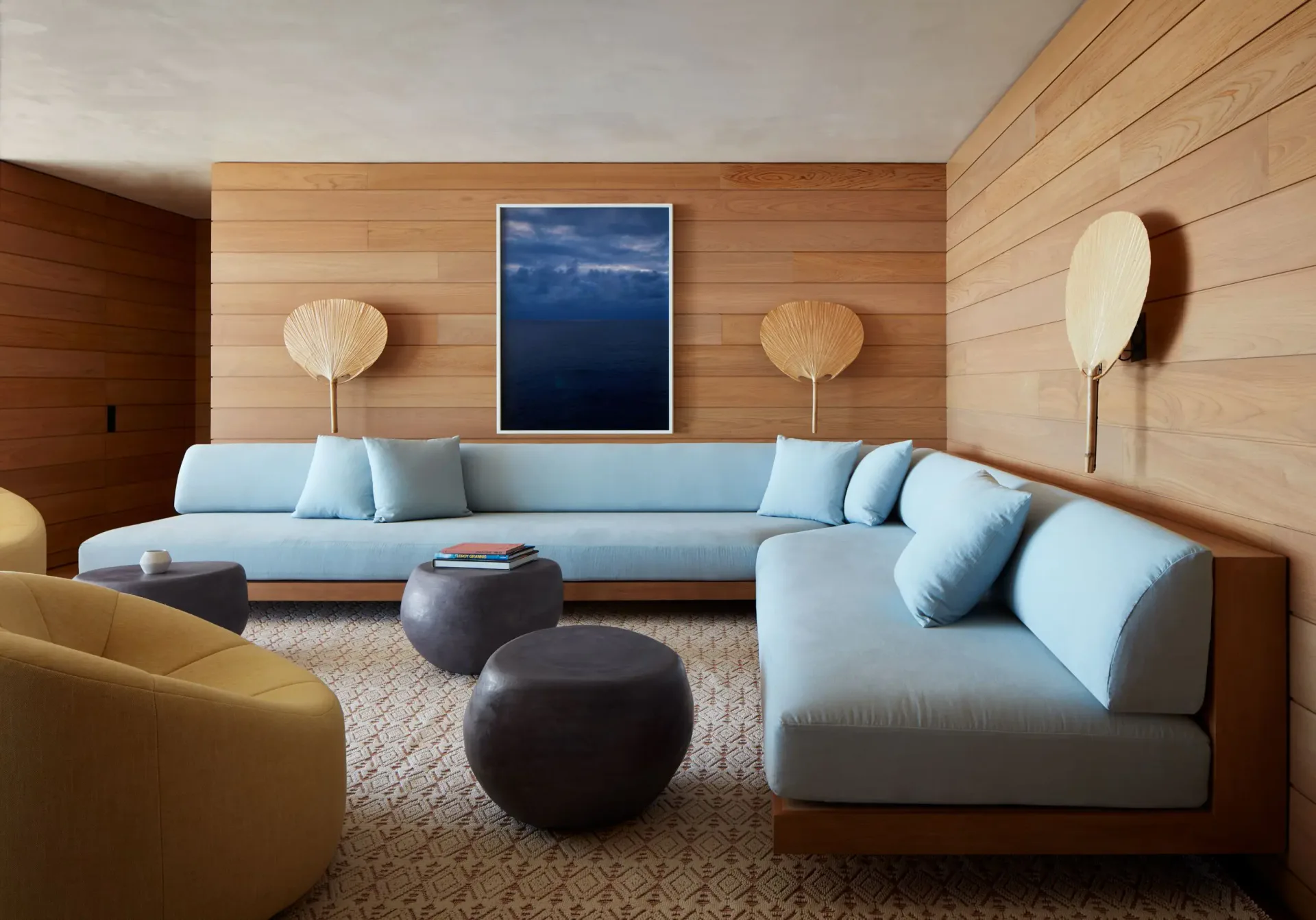 Modern living room with light blue sectional sofa, wooden wall paneling, dark poufs, and pendant lighting by Billy Cotton.