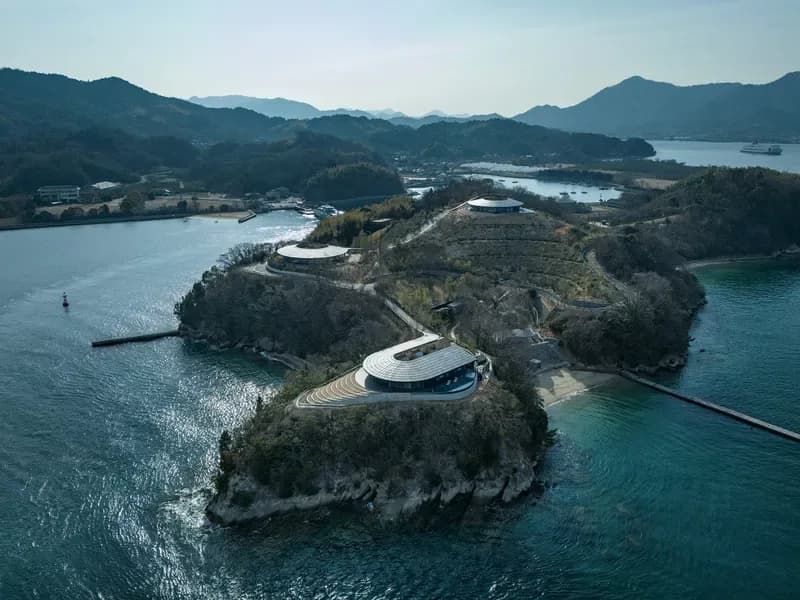 Aerial view of a futuristic curved white building on a forested peninsula surrounded by turquoise water and mountains, designed by Bjarke Ingels.