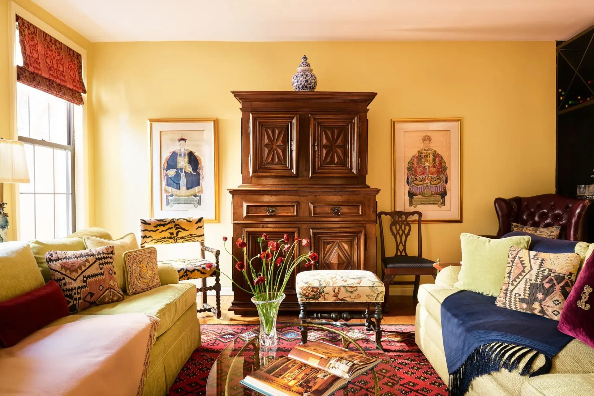 Yellow living room with dark wood cabinet, colorful patterned textiles, framed portraits, and layered furnishings by Blake Funston.