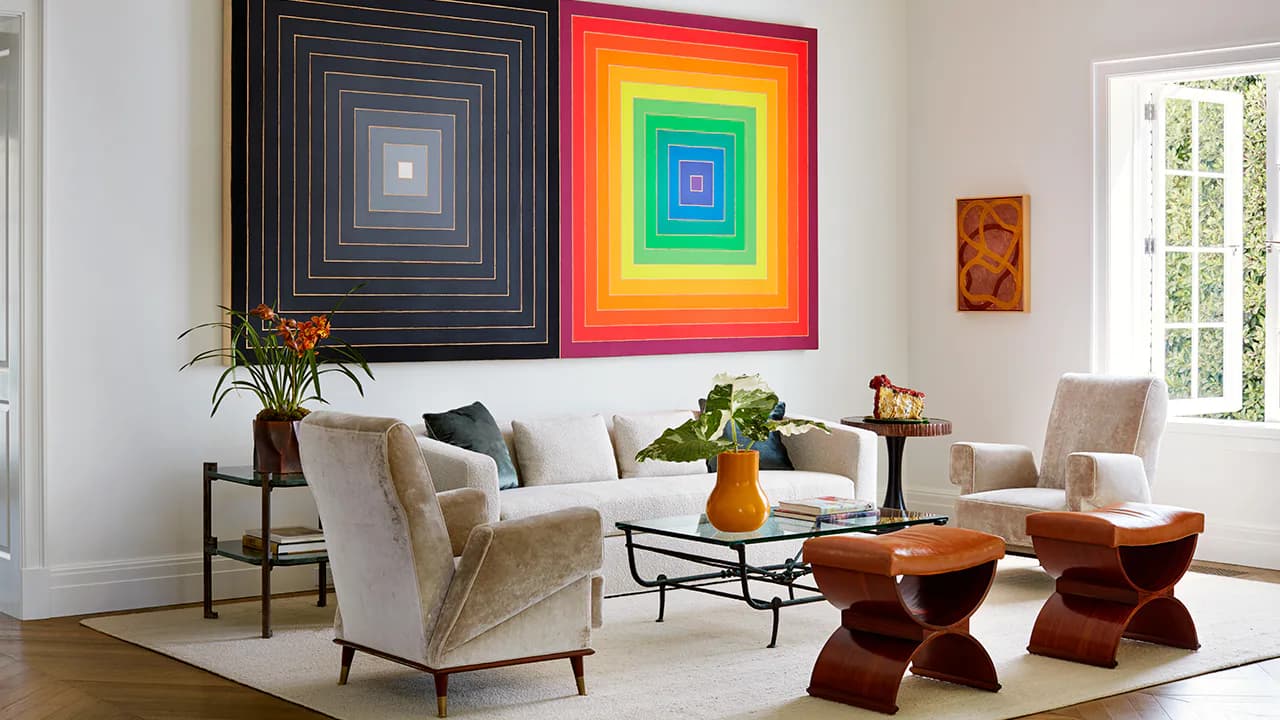 Bright modern living room with cream upholstered chairs, wood stools, geometric framed artwork in neon colors, potted plants, and floor-to-ceiling windows by Blue Williger.