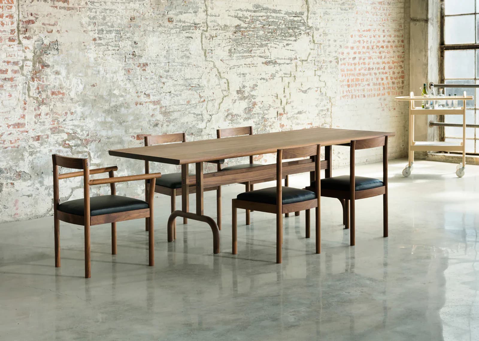 Mid-century modern dining set with walnut table and chairs featuring black upholstered seats in minimalist interior with exposed brick wall, designed by Bowen Liu.