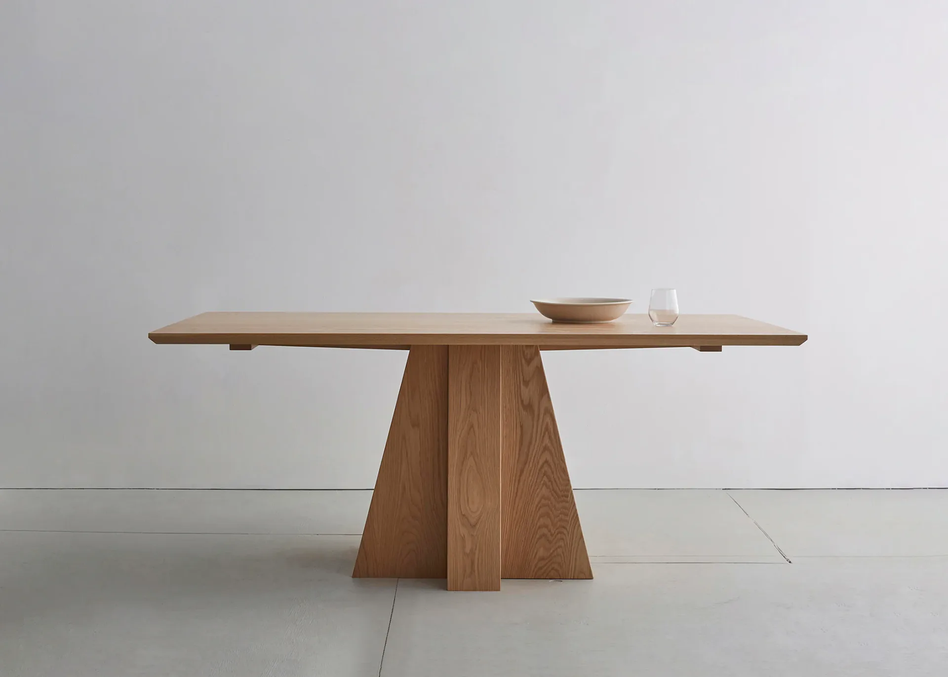 Minimalist wooden dining table with trapezoid pedestal base and elongated top, styled with small ceramic objects, by Bowen Liu.