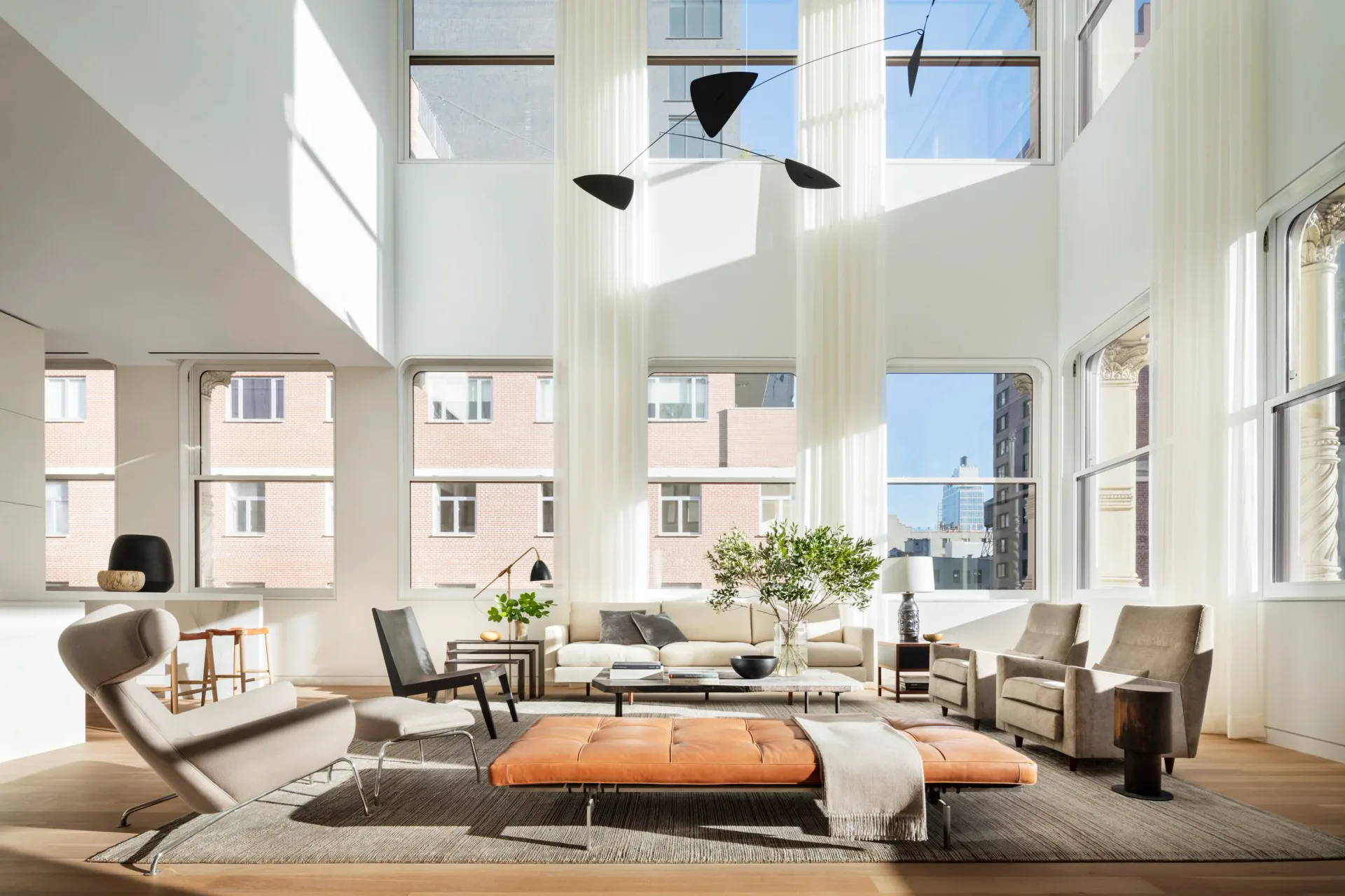 A bright, double-height loft living room with floor-to-ceiling windows, neutral palette, orange ottoman, and mid-century modern furniture by Brad Ford.