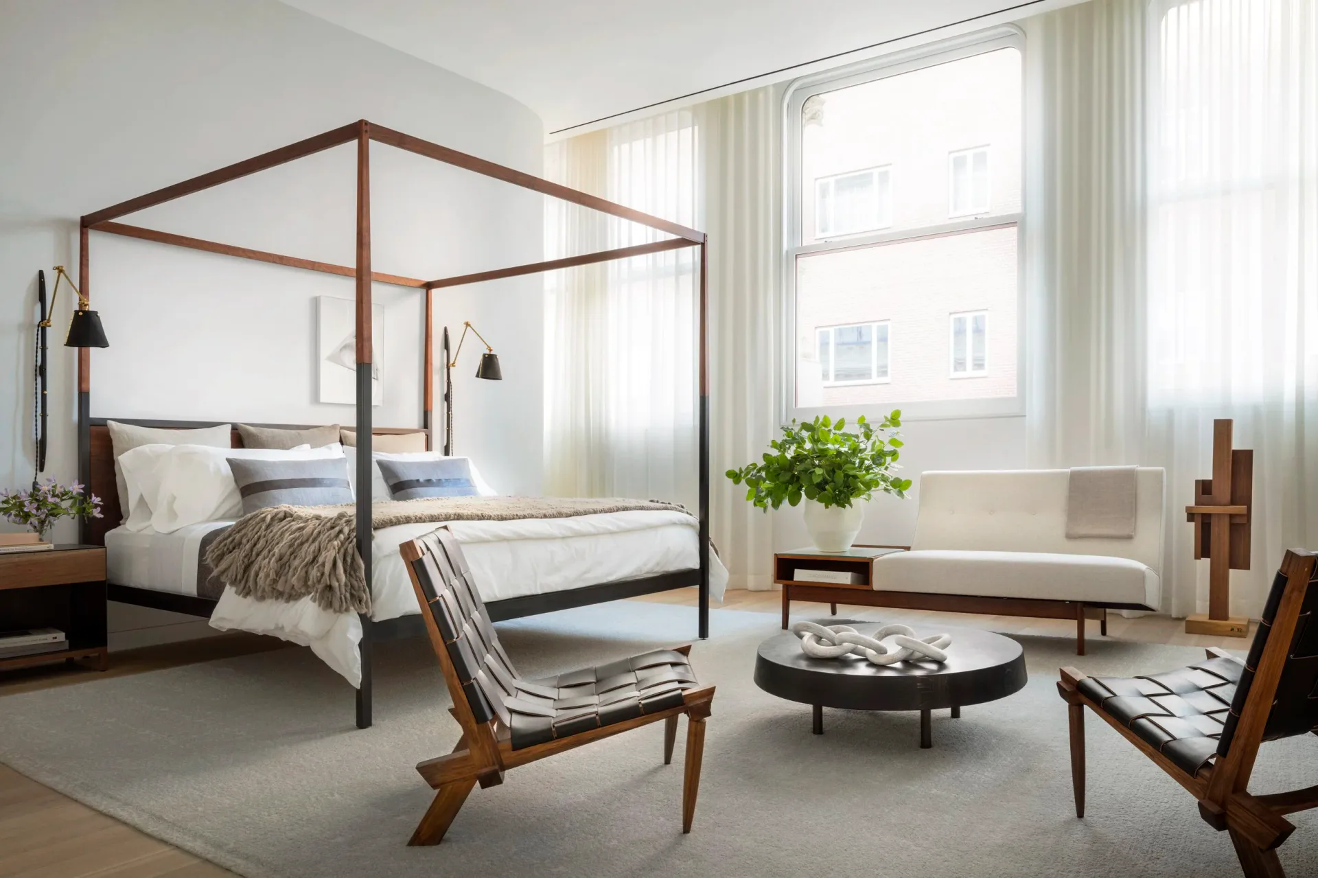 Bright contemporary bedroom featuring a wooden four-poster bed, mid-century modern seating, sculptural wood accents, and floor-to-ceiling windows with sheer curtains, designed by Brad Ford.