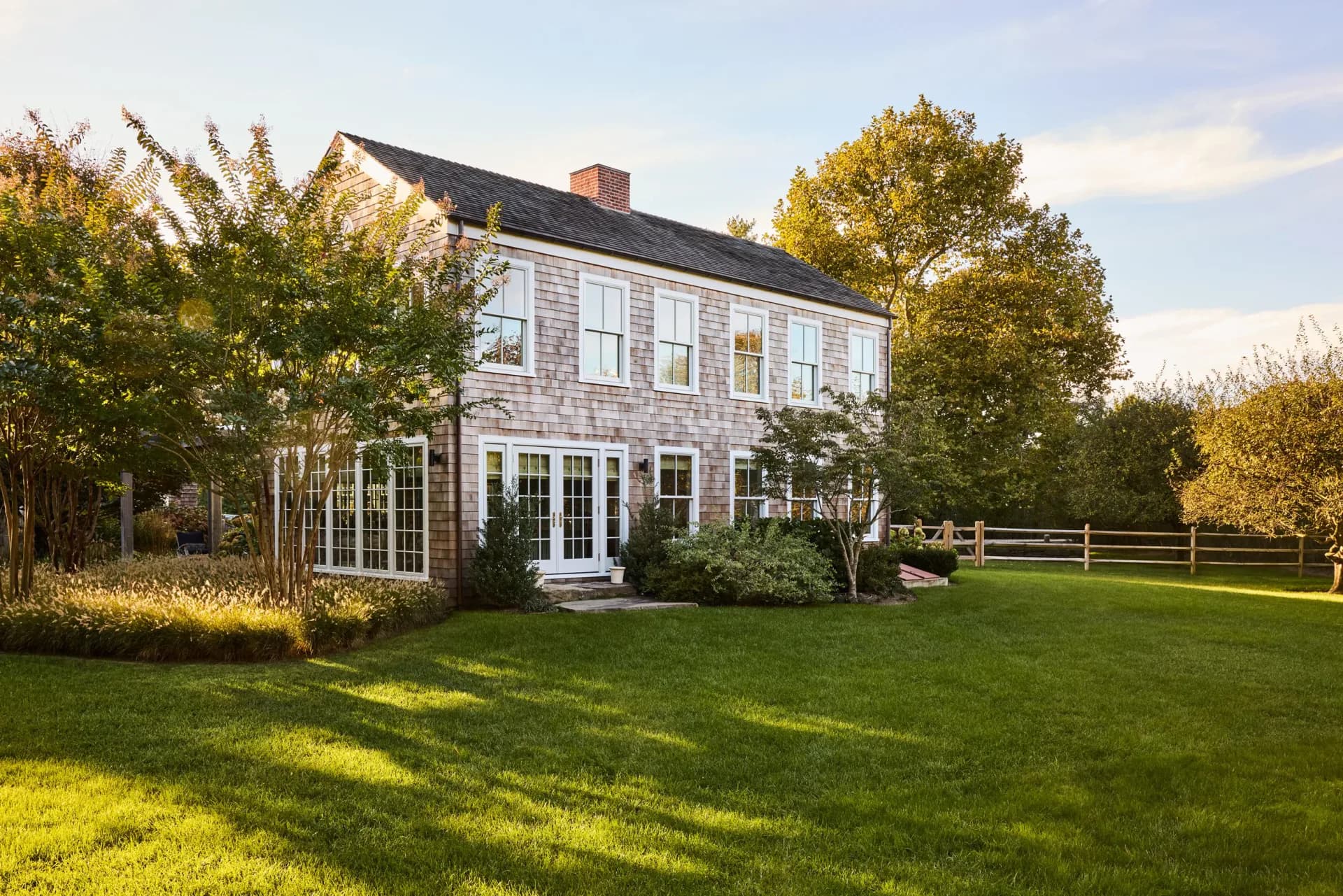 A classic New England farmhouse with shingle siding and white trim sits on manicured grounds surrounded by mature trees and a white fence by Bradley Silling.