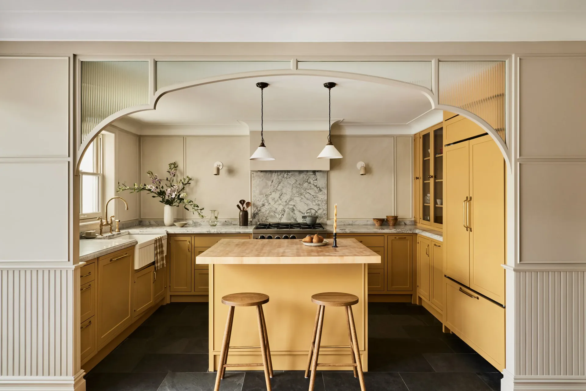 Warm-toned kitchen with golden wood cabinetry, marble backsplash, curved archway, island with wood stools, and dark flooring by Bradley Silling.