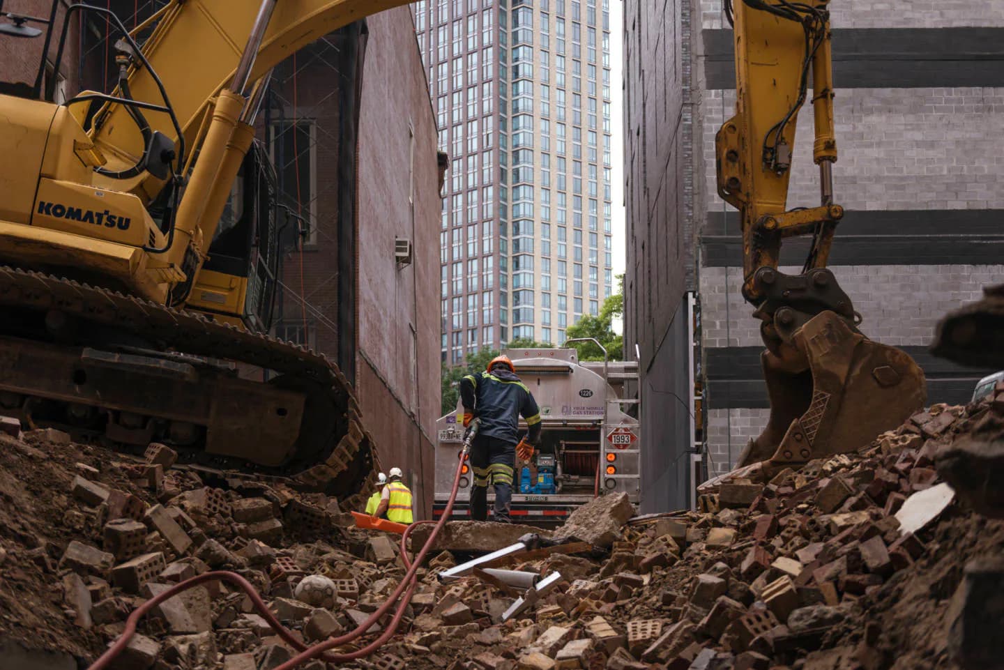 Urban excavation site showing heavy machinery and workers among demolished concrete and soil, with tall buildings visible in background by Brandon Lim.
