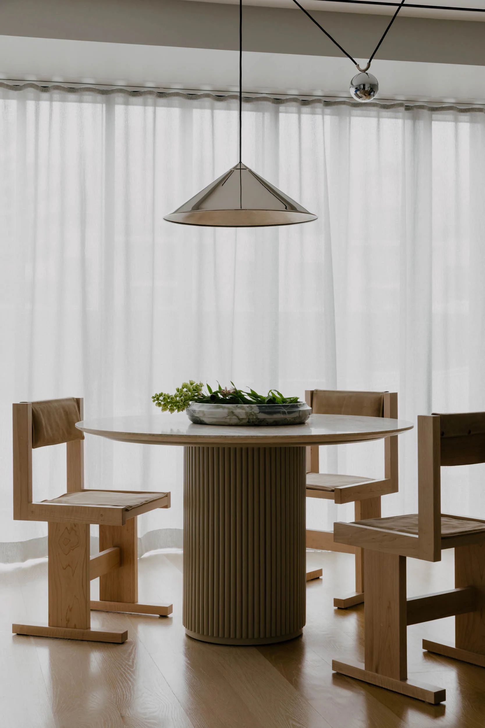 Minimalist dining space with round table on fluted pedestal base, wooden cantilever chairs, pendant light, and sheer curtains by Brendan Mahoney.
