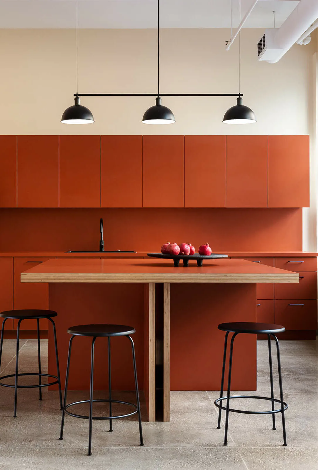 Minimalist kitchen with bold burnt-orange cabinetry, light wood island, black pendant lights, and red apples, designed by Brendan Mahoney.