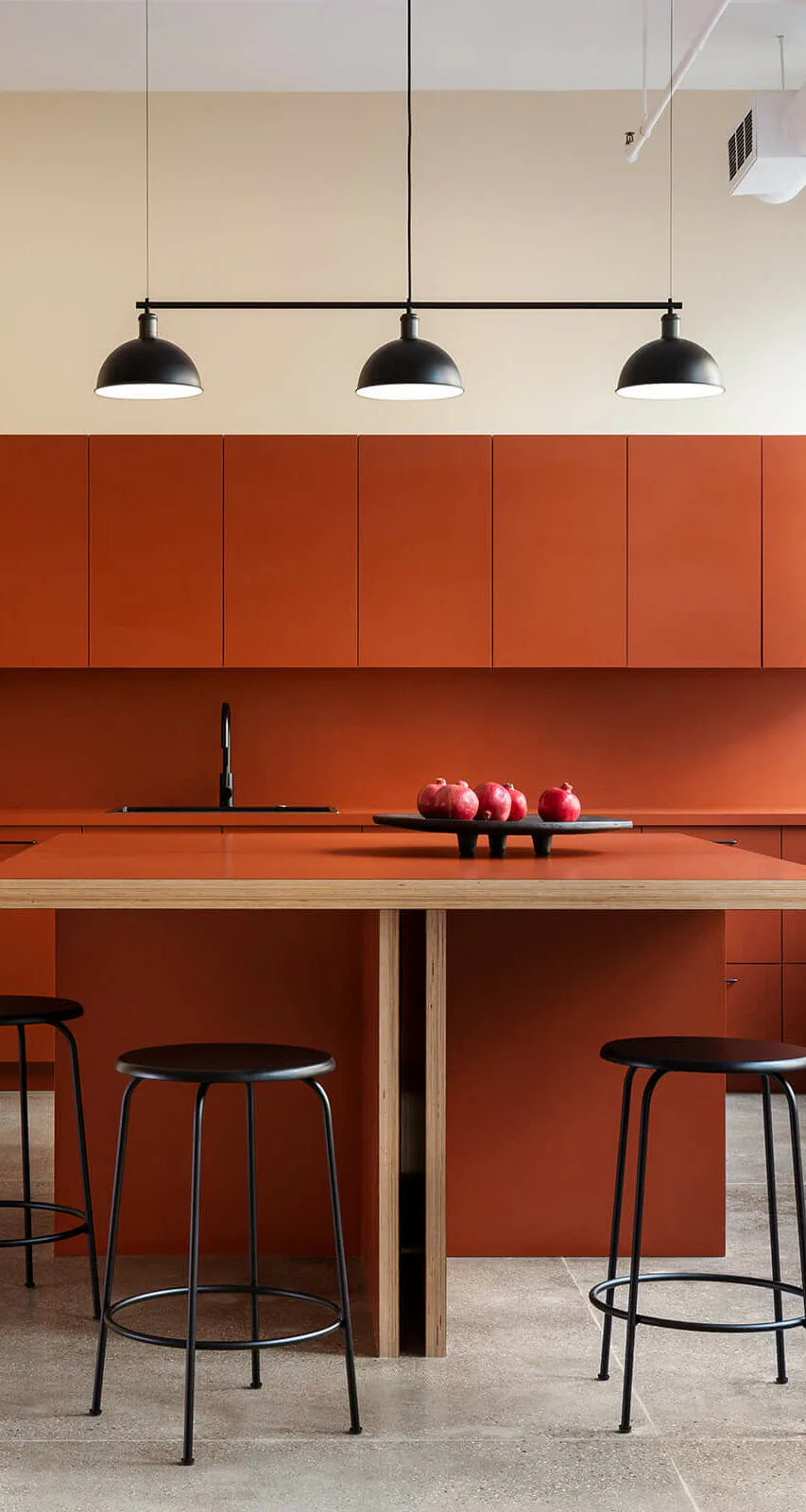 Modern kitchen with burnt-orange cabinetry and island, black pendant lights, natural wood countertop, bar seating, and apples on counter by Brendan Mahoney.