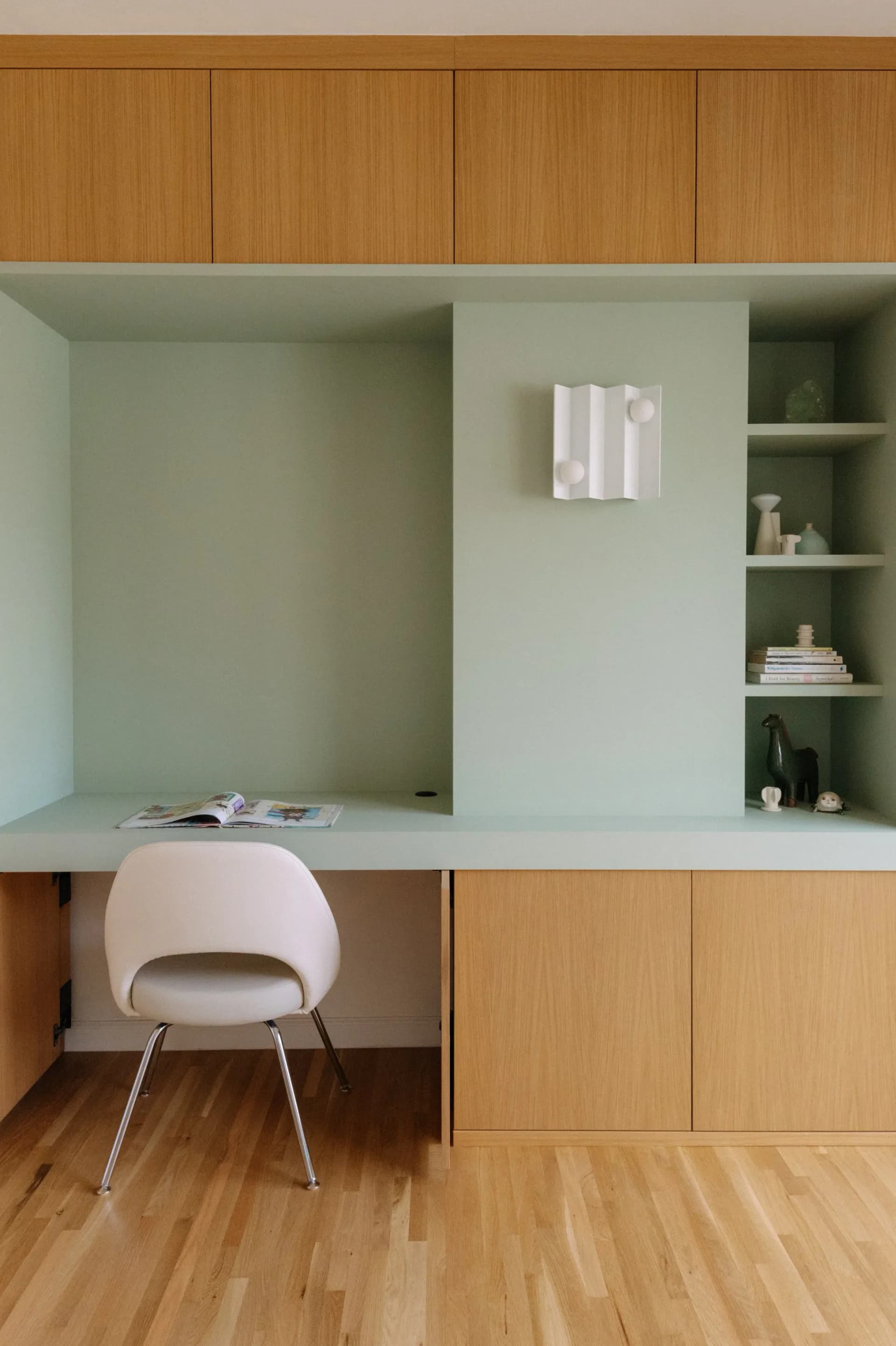 Modern home office with pale green built-in cabinetry, natural wood upper cabinets, white desk, floating shelves, and warm wood flooring by Brenna Thompson.