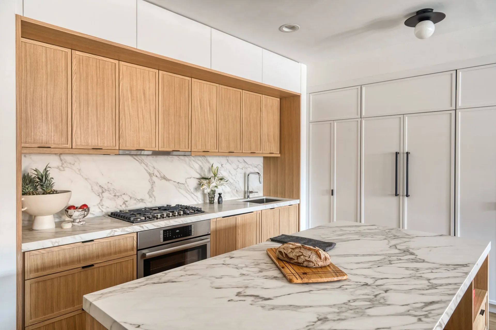 Modern kitchen with warm wood cabinetry, marble countertops, and white upper cabinets by Brenna Thompson.