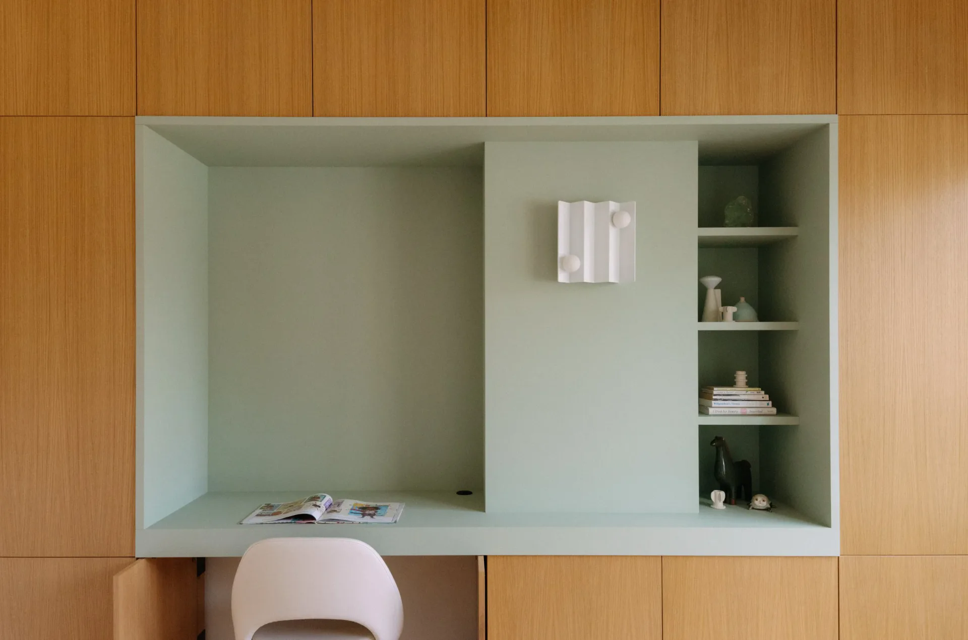 Modern built-in desk nook with soft sage green interior, pale pink chair, natural wood cabinetry, and integrated shelving by Brenna Thompson.