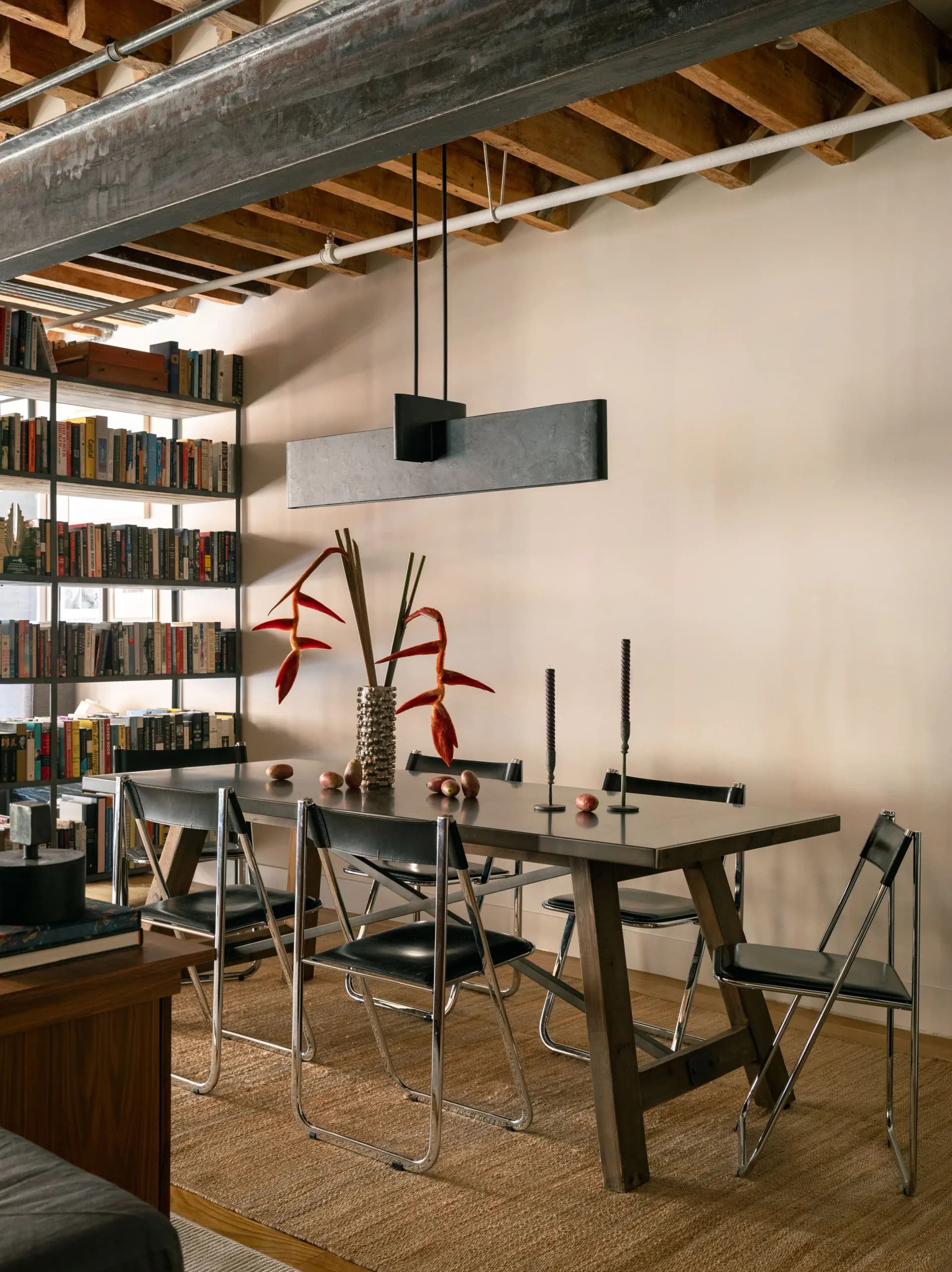 Modern dining room with dark wood table, metal chairs, exposed beam ceiling, built-in bookshelves, pendant light fixture, and red floral arrangement by Brian Cash.