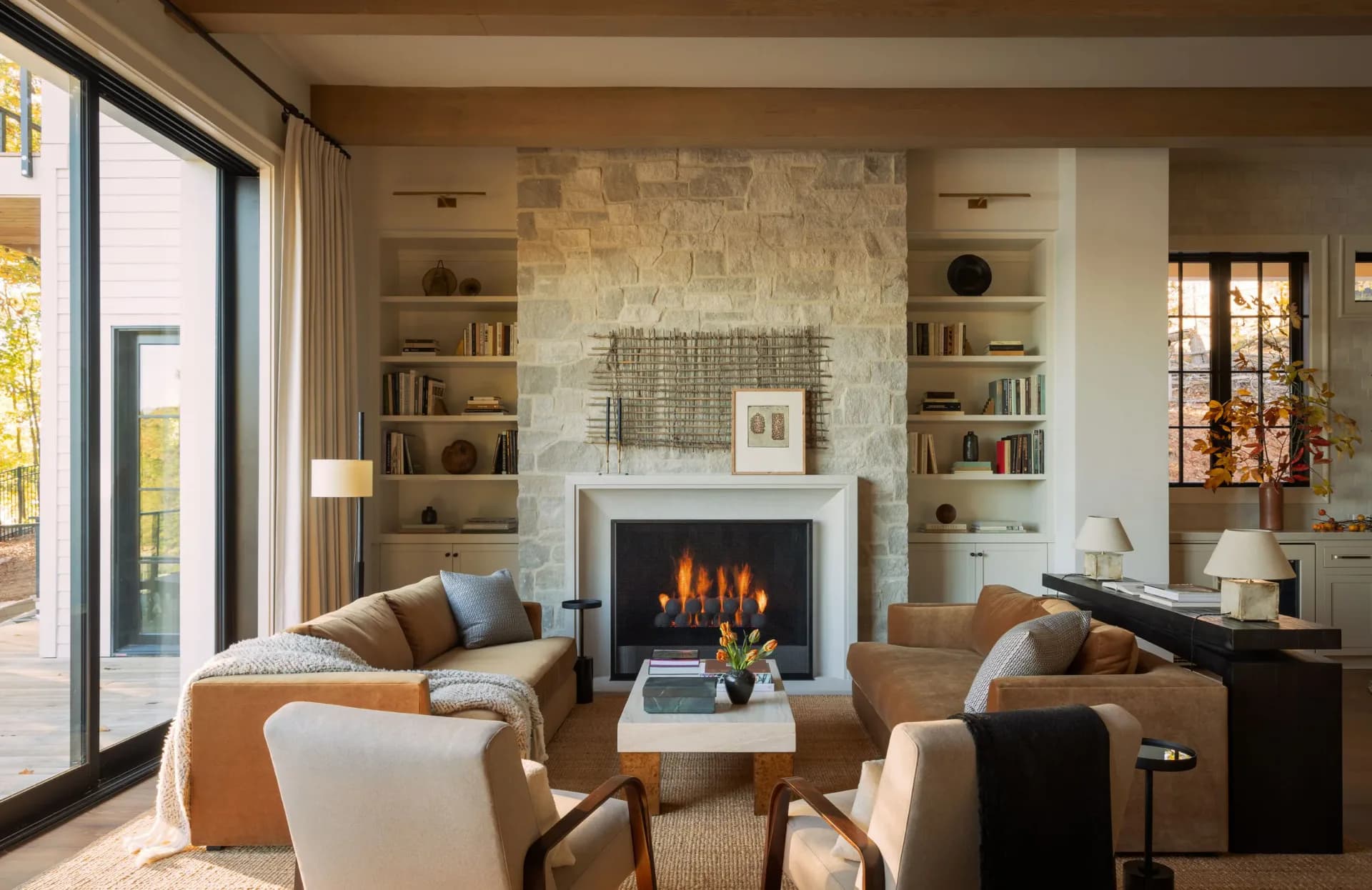 Warm contemporary living room with stone fireplace, built-in shelving, cream armchairs, and large glass doors opening to exterior, designed by Brian Cash.