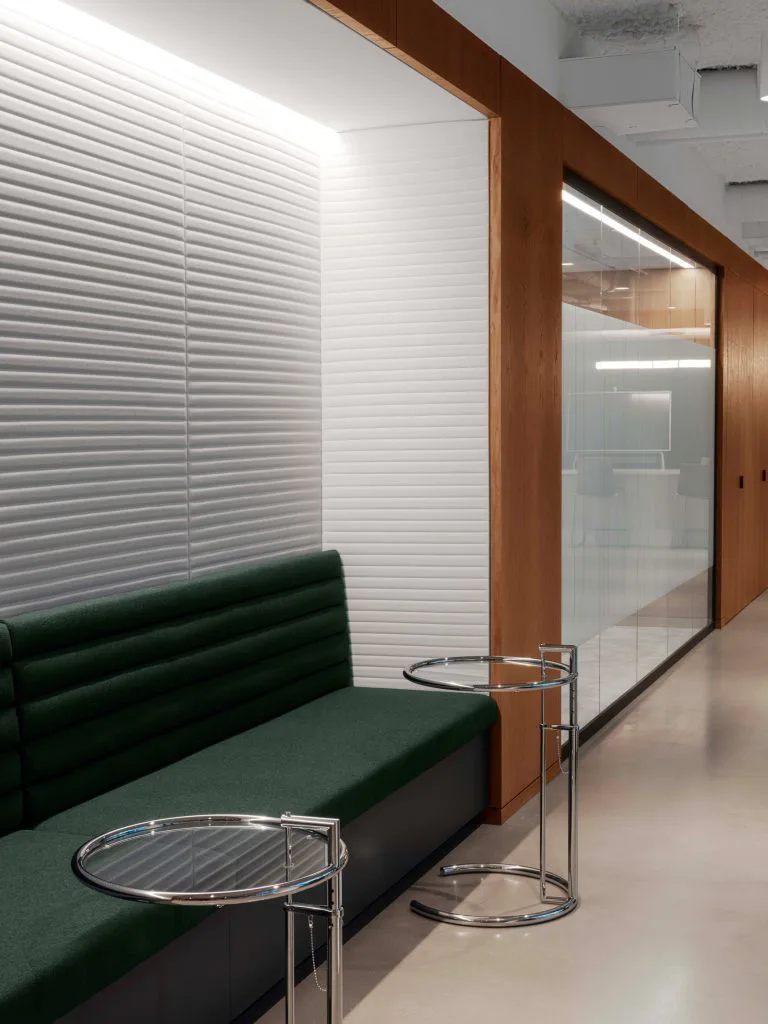 Modern office waiting area featuring a dark green upholstered bench, frosted glass partition wall, horizontal white louvers, and minimalist chrome side tables by Brian Orter.