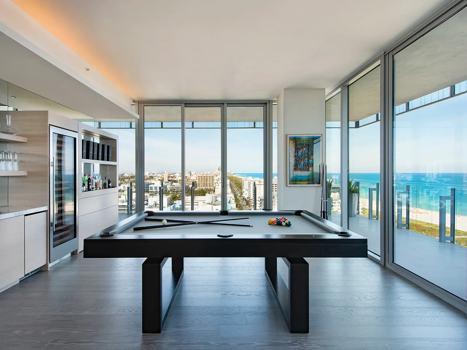 Modern luxury penthouse with black pool table, floor-to-ceiling windows overlooking coastal views, minimalist white cabinetry, and polished concrete flooring by Briana Kimball.