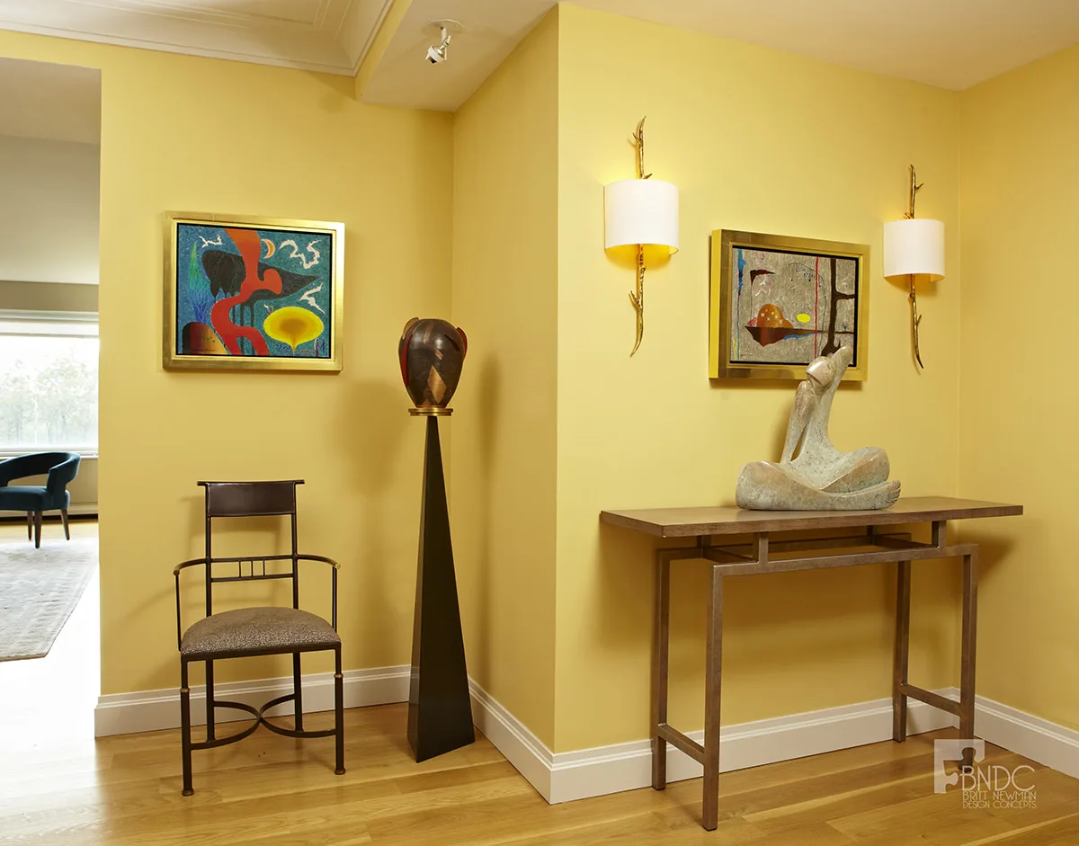 Yellow gallery room with wooden console table, sculptural bust, framed artwork, pendant lights, and modern furniture by Britt Newman.