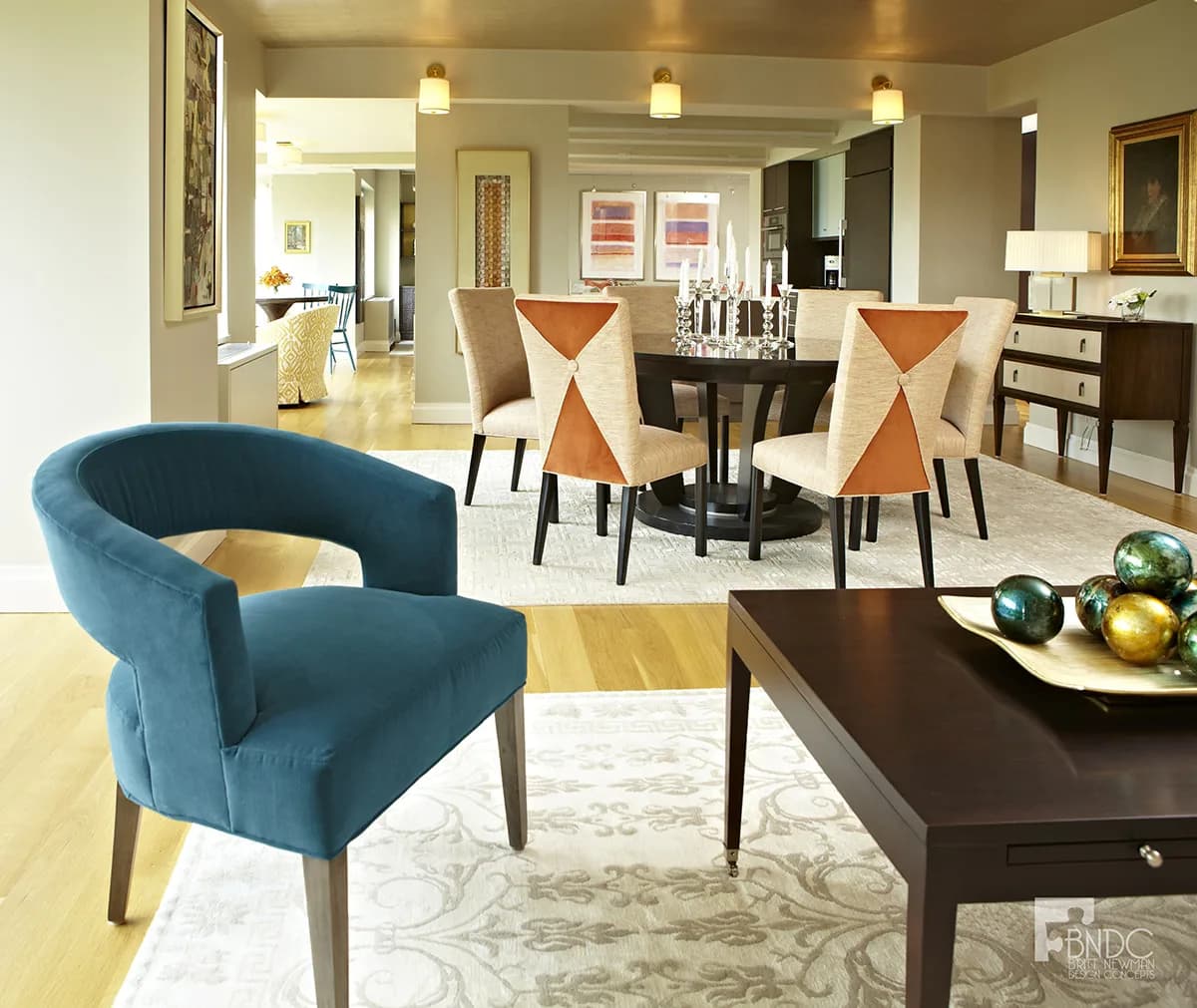 Open-concept living and dining space featuring a teal curved armchair, dark round dining table with orange X-back chairs, and cream-toned finishes by Britt Newman.
