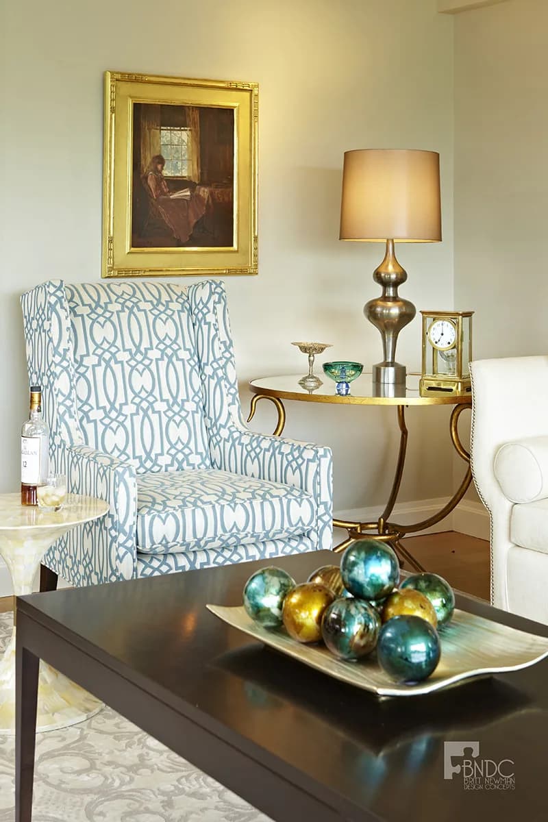 Elegant living room interior featuring a blue and white patterned wingback chair, brass table lamp, framed portrait, and decorative brass spheres on dark coffee table by Britt Newman.