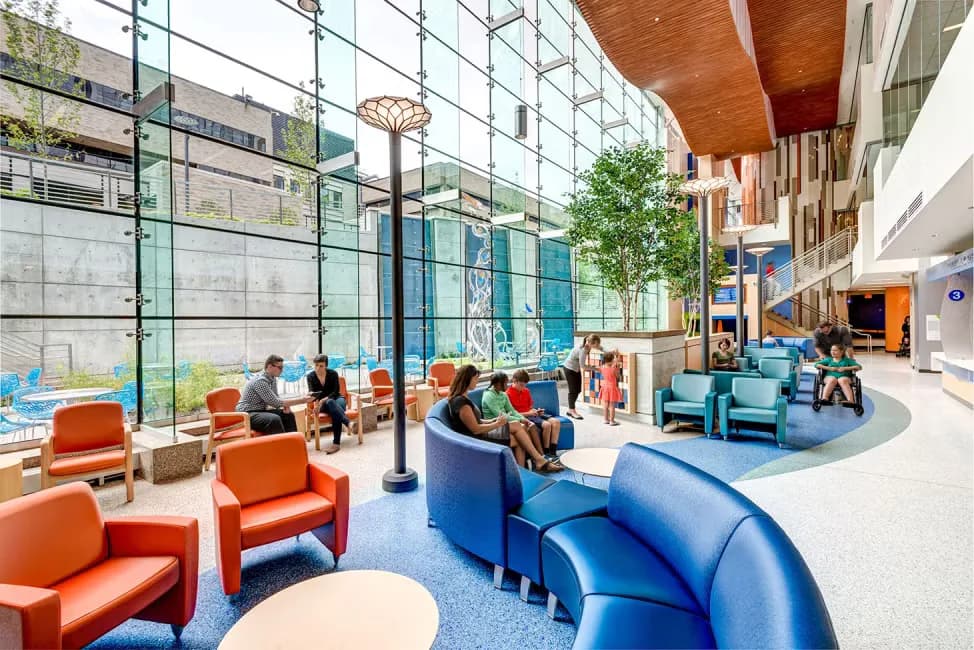 Modern institutional atrium with orange and blue seating, blue carpet, wooden ceiling elements, large windows, and greenery by Brooke Horan.