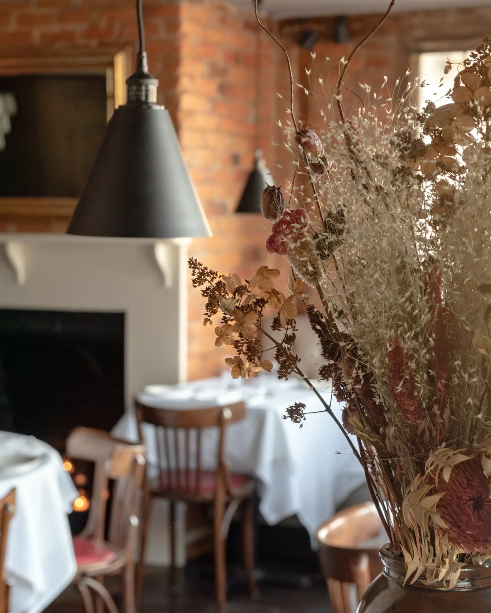 Warm restaurant interior featuring dried floral arrangements in glass vases, pendant lighting, exposed brick walls, and white tablecloths by Brooke Lane.