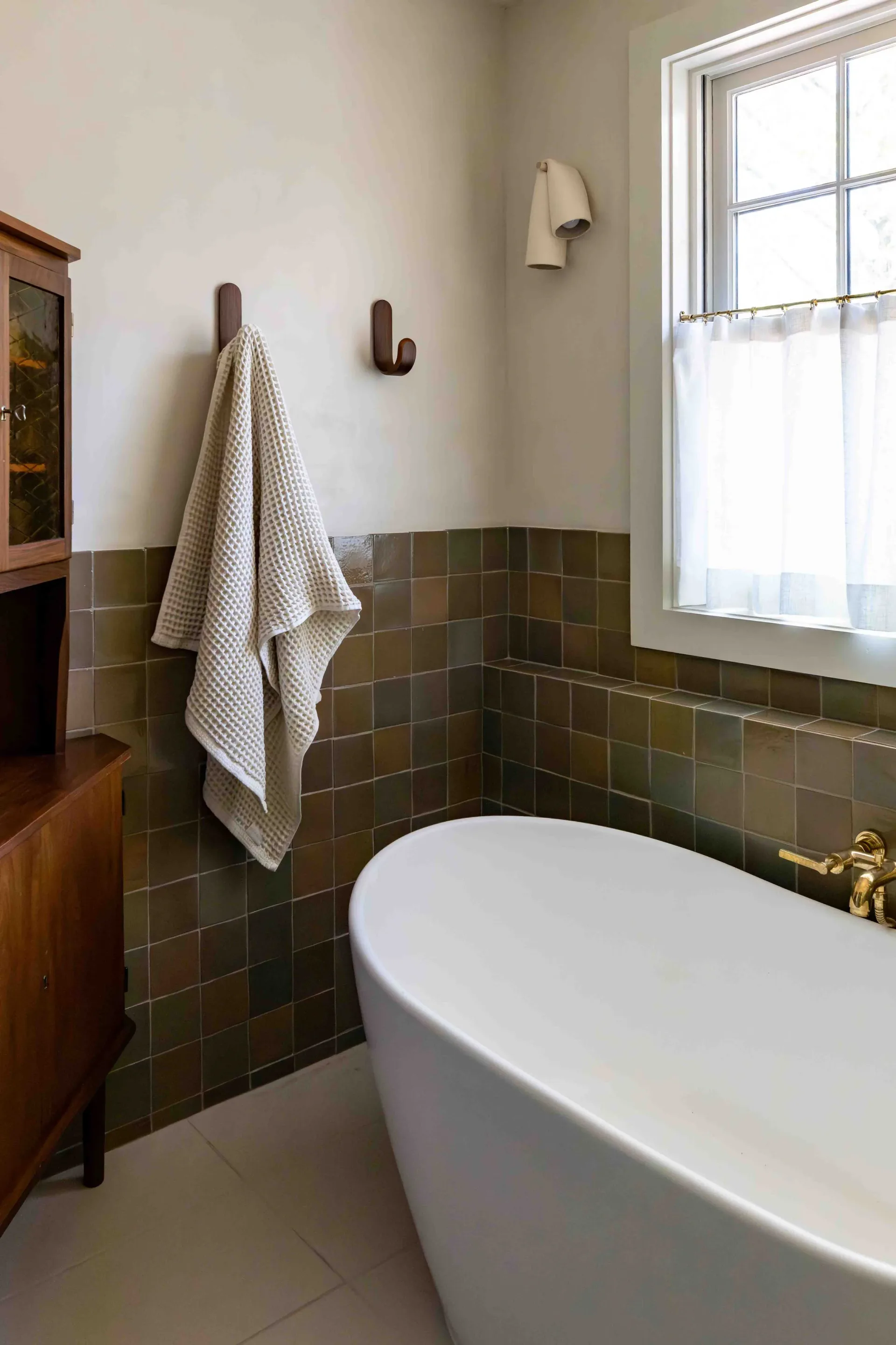 Modern bathroom with freestanding white tub, olive-green tile wainscoting, wooden vanity, and natural light from large window by Brooke Lane.