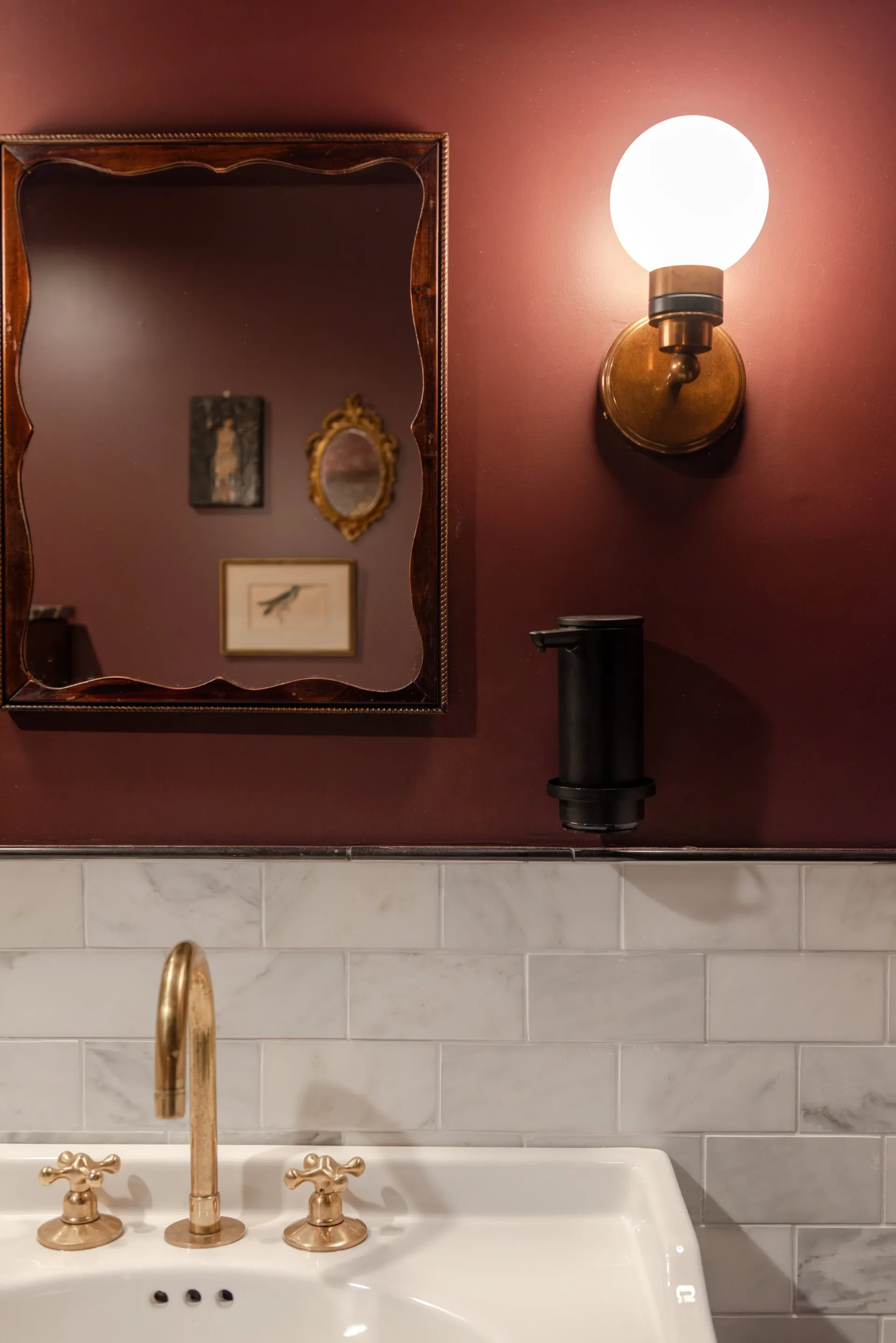 Luxurious powder room with deep mauve walls, white subway tile, brass fixtures, and vintage mirror with sconces by Brooke Lane.