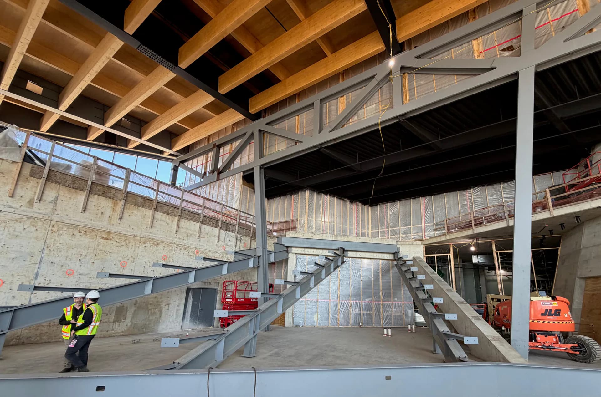 Multi-level industrial construction site with exposed concrete floors, steel beams, and wooden ceiling structure, featuring a worker in safety gear by Bryan Dorsey.