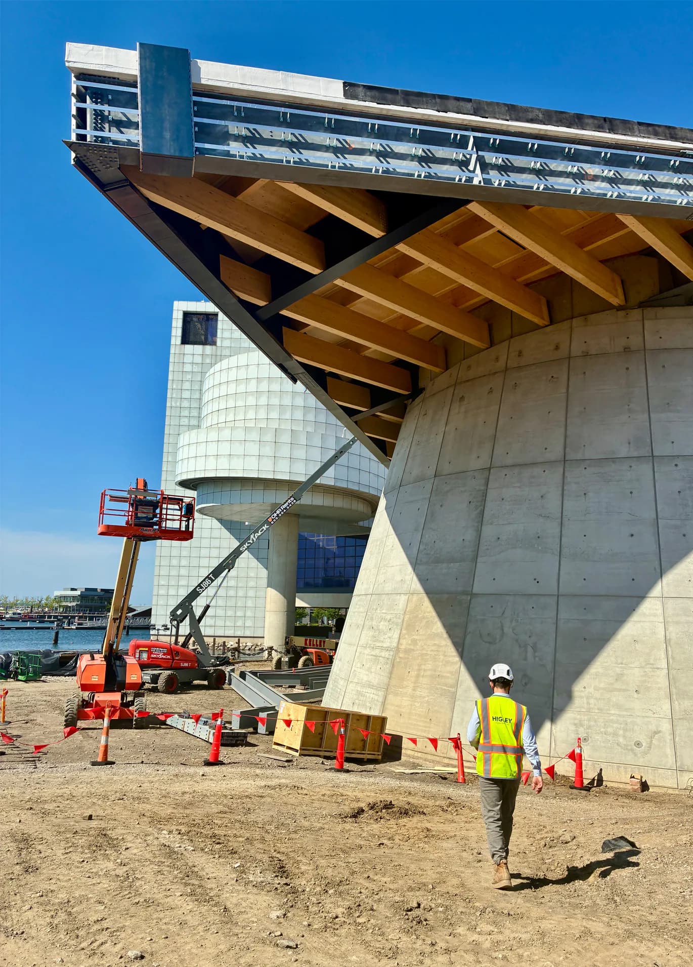 Concrete brutalist building with dramatic cantilevered wooden beam structure under construction, featuring exposed wood joinery and metal framework against clear blue sky by Bryan Dorsey.