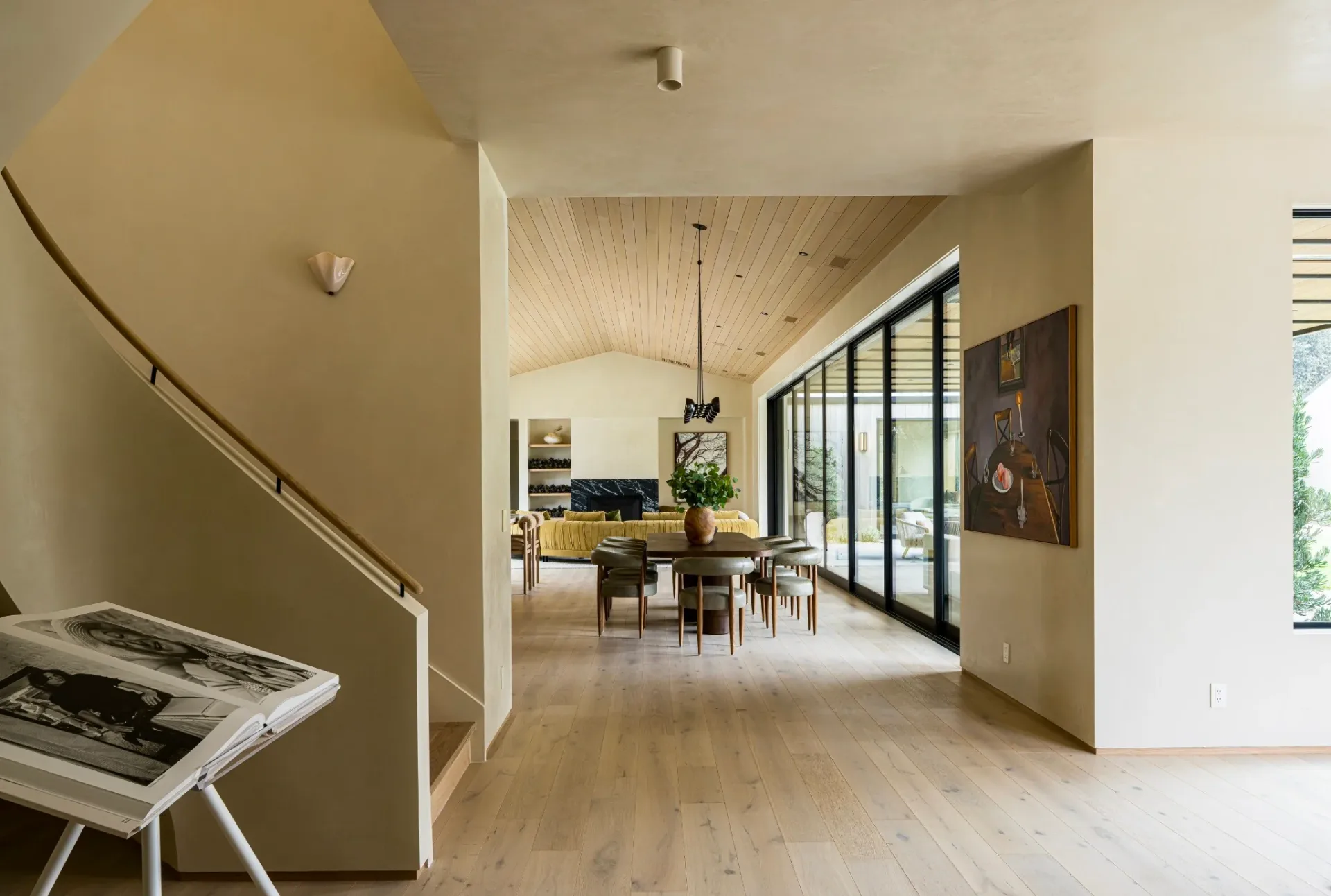 Light-filled open-plan interior with natural wood flooring, white walls, and floor-to-ceiling black-framed windows, featuring a workspace and dining area by Burcu K.