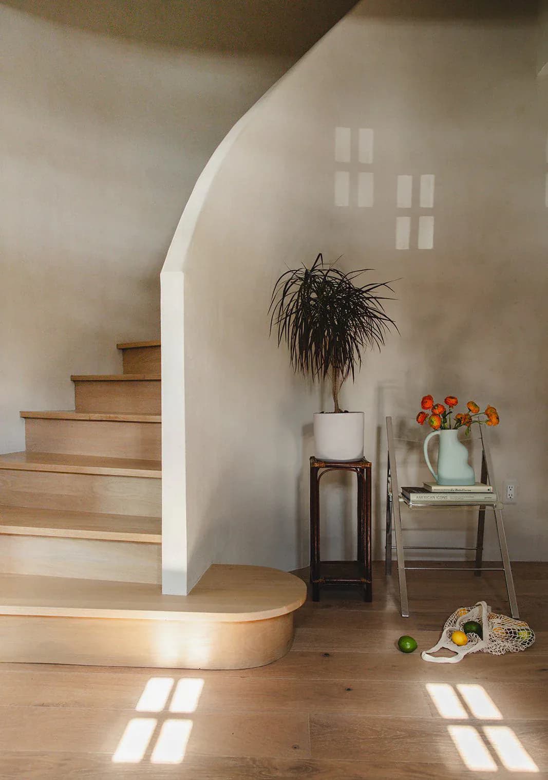 Minimalist interior with curved white walls, wooden stairs, and natural light casting geometric shadows on light wood flooring, featuring potted plants and sculptural furnishings by Burcu K.