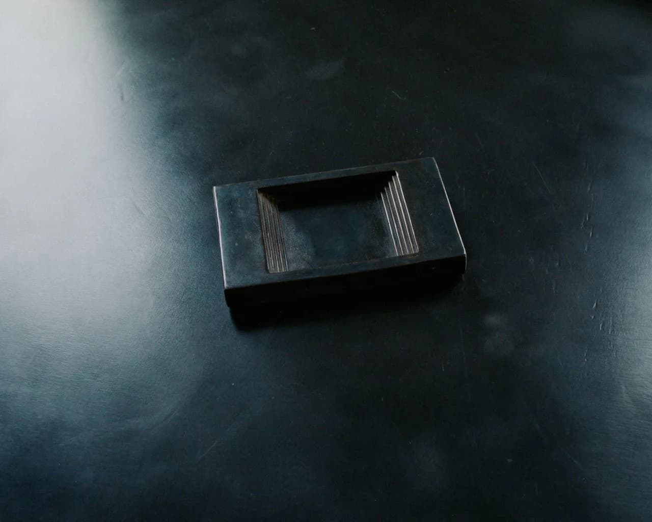 Dark metallic rectangular object with geometric cutout detail photographed on textured surface, minimalist product design by Cal Summers.