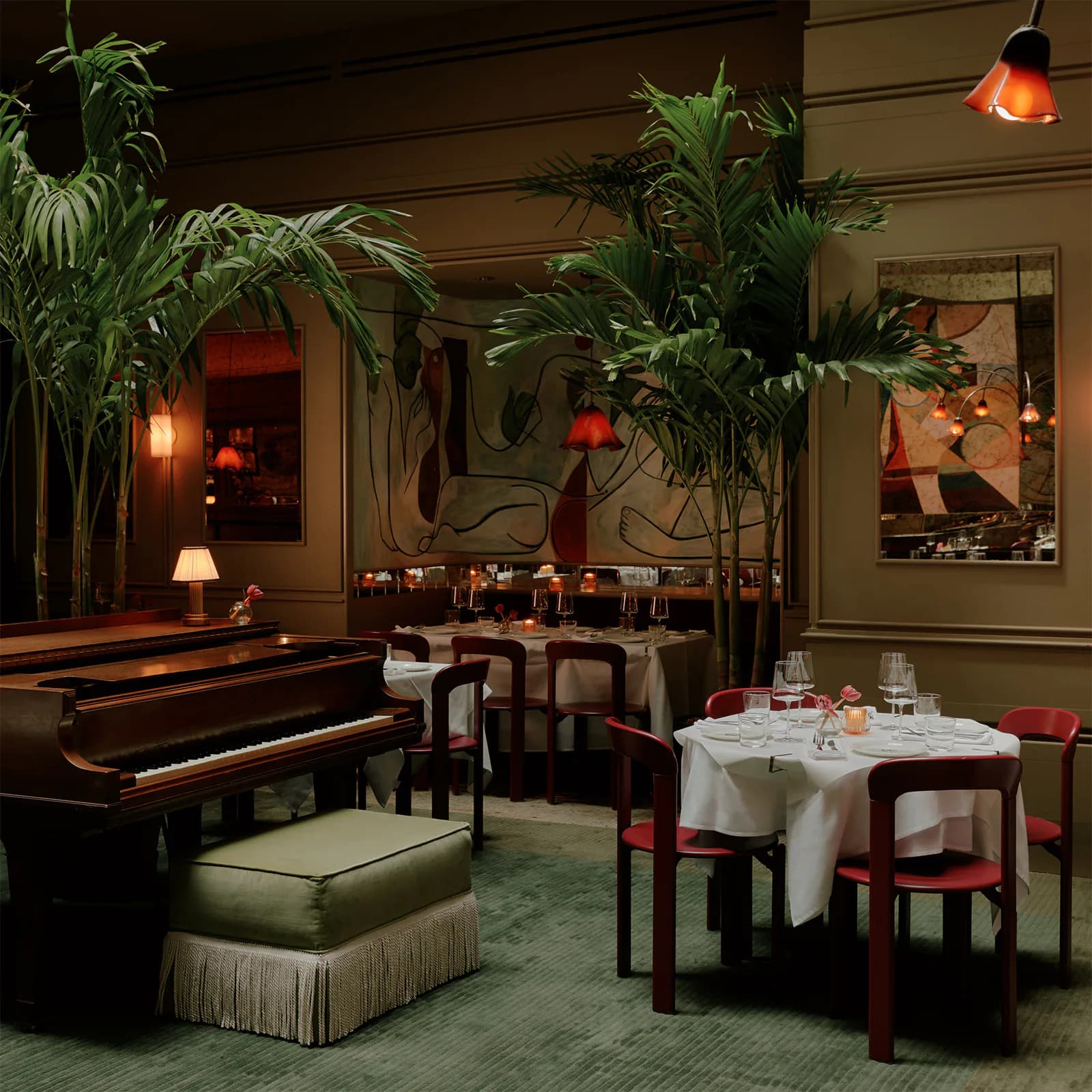 Elegant dining room with grand piano, lush tropical plants, warm amber lighting, white tablecloth dining table with red chairs, and rich wood paneling by Callie Forsythe.