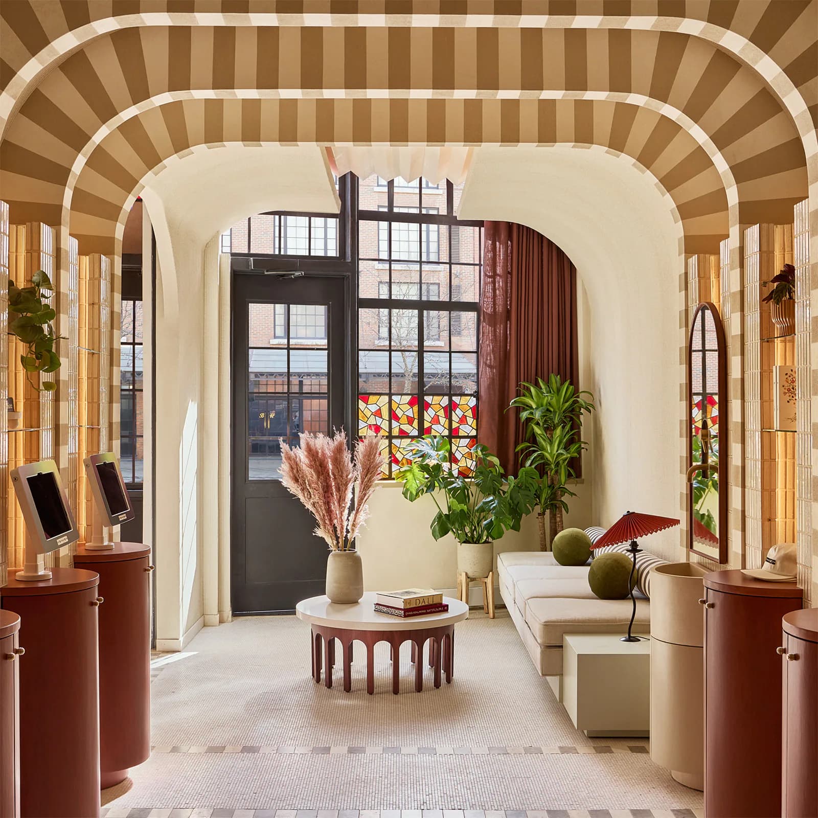 A sunlit residential gallery space features cream walls, striped barrel-vault ceilings, terracotta cylindrical furnishings, and large black-framed windows with burgundy curtains, displaying curated plants and minimalist décor by Callie Forsythe.