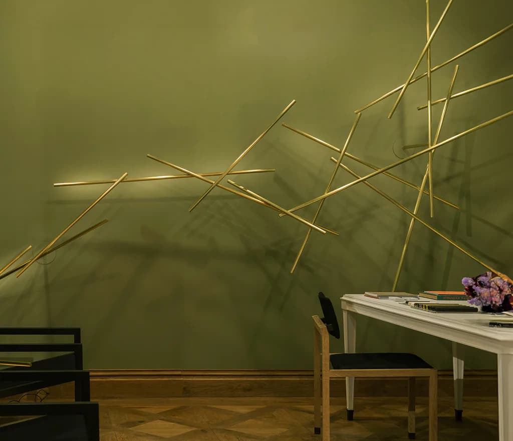 Minimalist interior with sage green wall featuring dynamic brass linear installation art, wooden flooring, and modern furniture by Cameron Crockford.