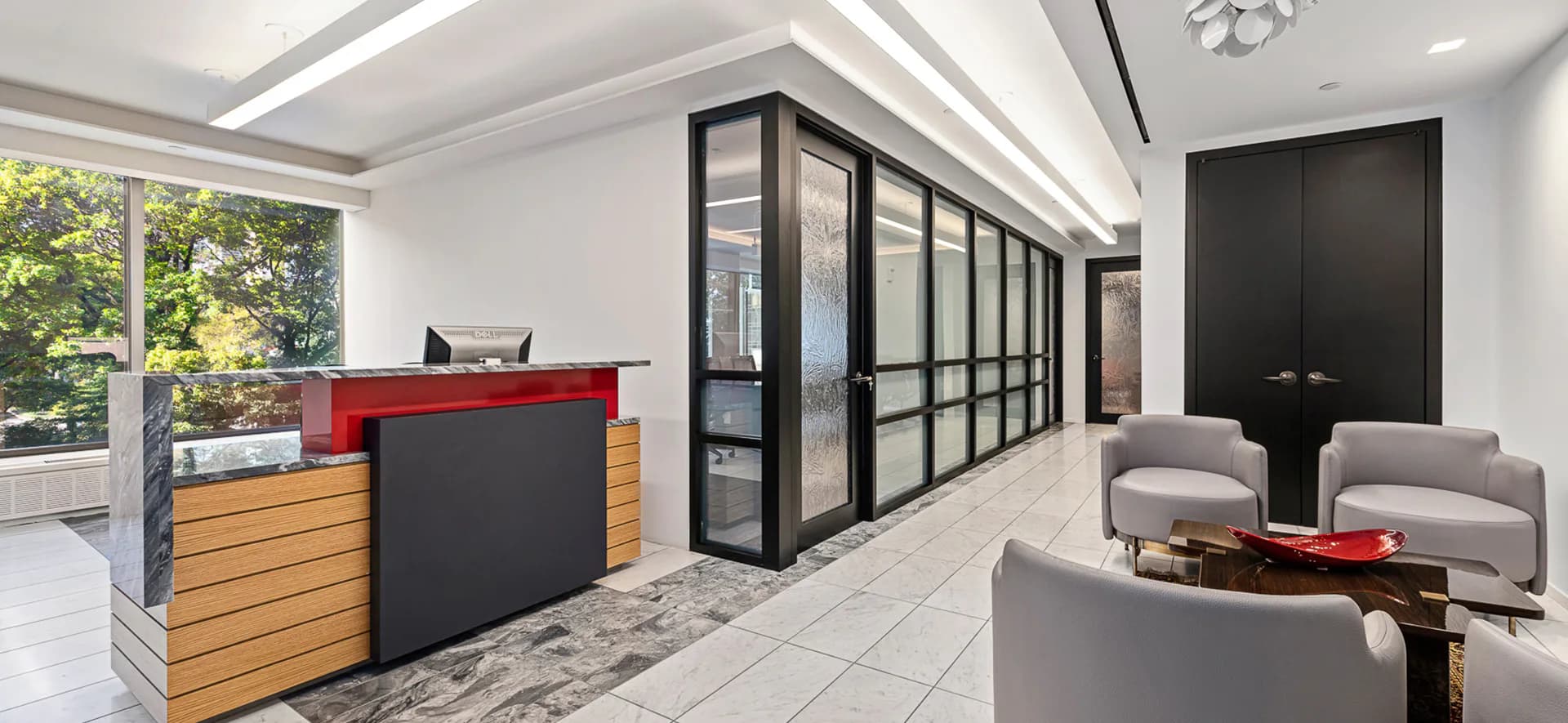 Modern office reception area with wood and black metal desk, glass partition walls, gray seating, and polished tile flooring by Carmelo Mazzaferro.