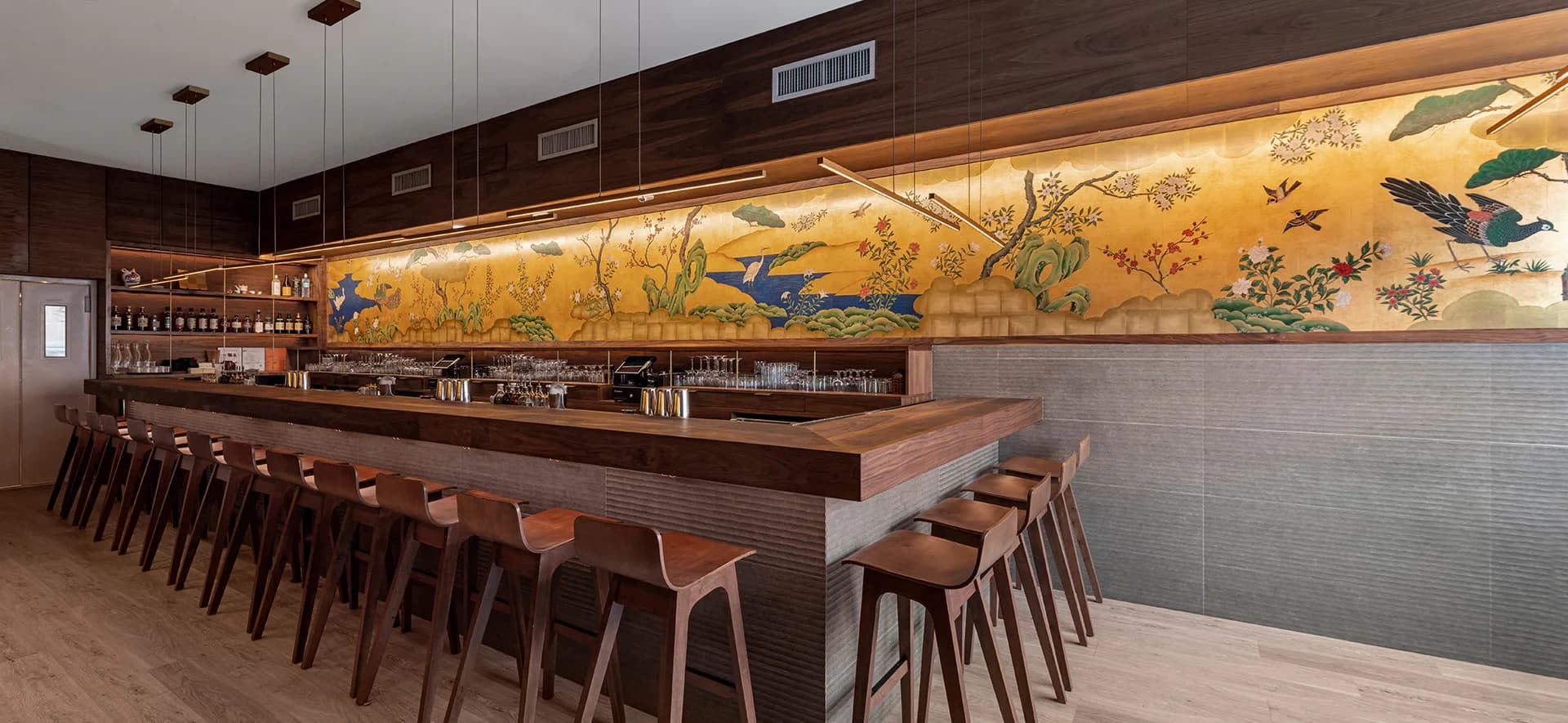 Modern bar interior featuring a long wooden counter with matching stools and a striking golden mural with birds and natural elements by Carmelo Mazzaferro.