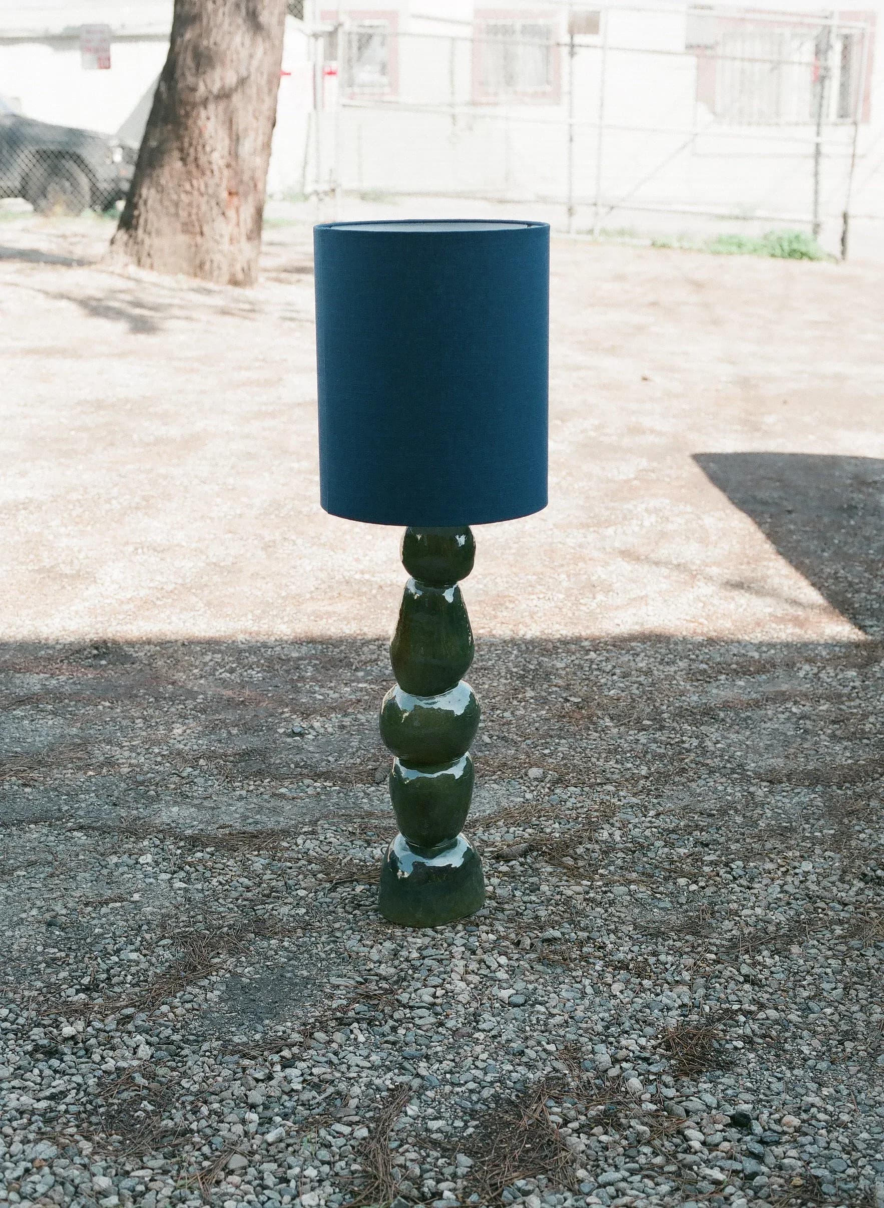 Dark green ceramic stacked-sphere table lamp with teal cylindrical shade, photographed outdoors on gravel by Carmen D'Apollonio.