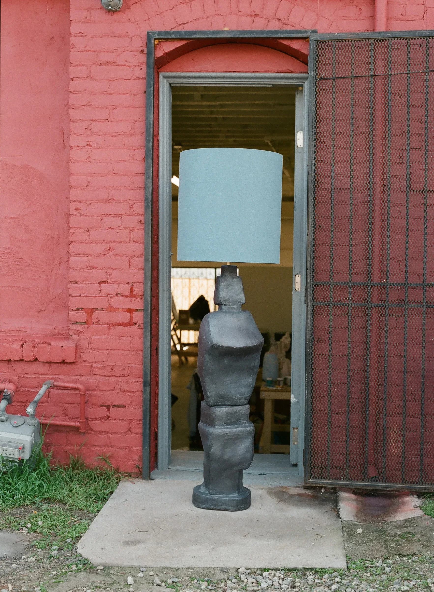 A dark sculptural torso installation in a coral-pink brick gallery space with industrial metal door frame by Carmen D'Apollonio.