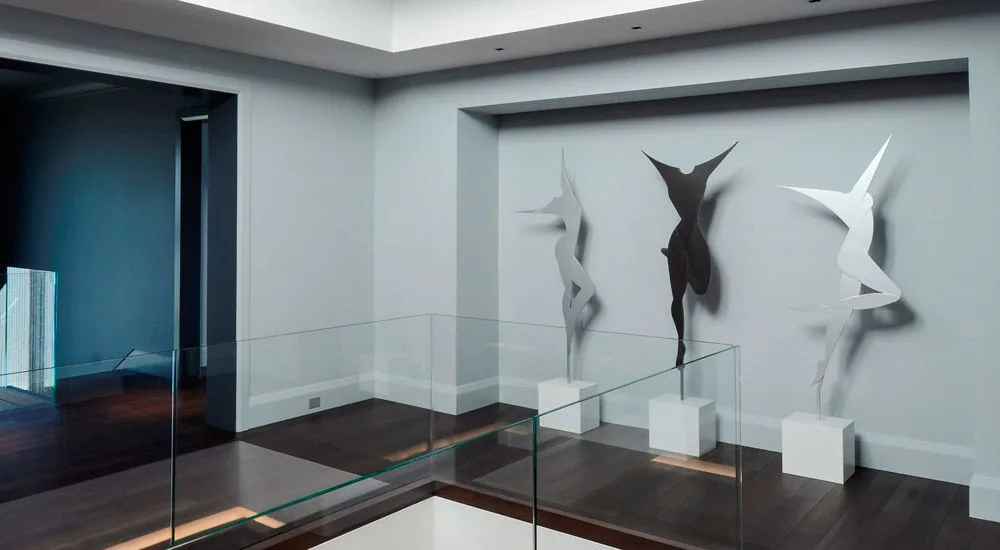 Modern gallery interior with glass display case showcasing three white sculptural forms against a light gray wall, minimalist design by Carol Brewer.