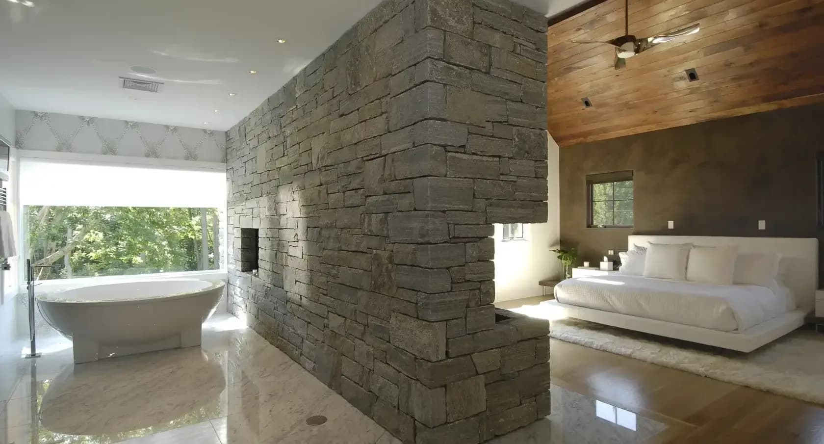 Luxury master suite with stone accent wall, freestanding sofa and integrated spa bathroom with marble floors by Carol Kurth.
