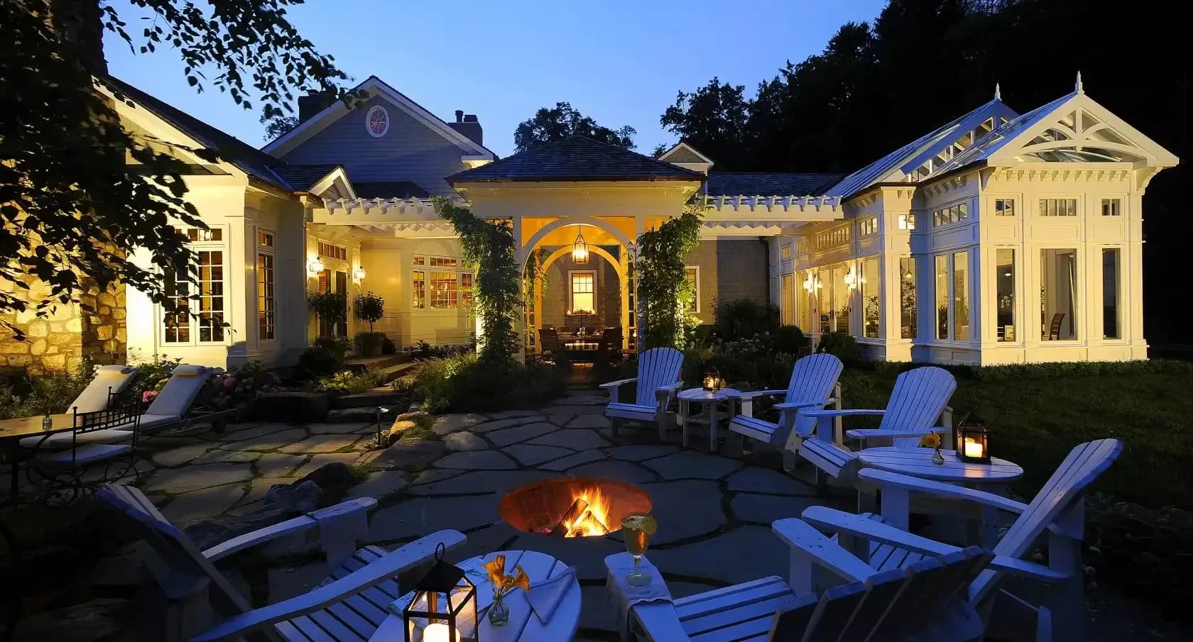 Illuminated residential courtyard with yellow pavilions, blue Adirondack chairs, and central fireplace at dusk, designed by Carol Kurth.