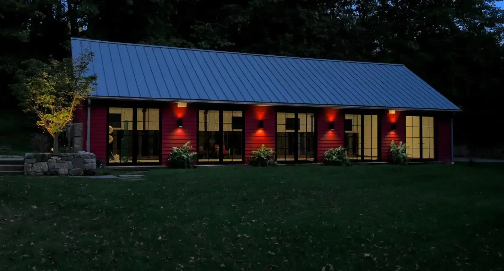 Modern residential barn with deep red exterior, blue metal roof, and warm interior lighting glowing through large windows, set in evening landscape by Carol Kurth.