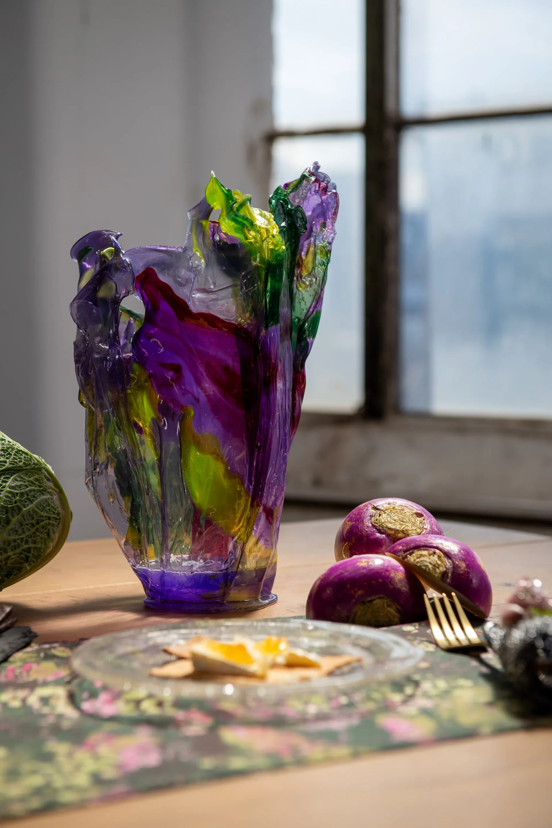 Sculptural glass vessel in purple and chartreuse with organic forms, displayed on wooden table with decorative objects and city view, by Caroline Zimbalist.