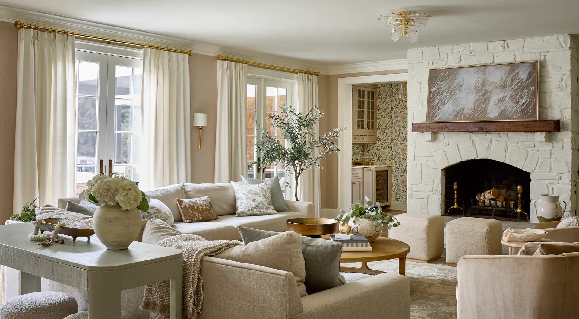 Elegant living room with cream and neutral tones, white brick fireplace, large windows, upholstered seating, and refined furnishings by Caroline Zogheb.