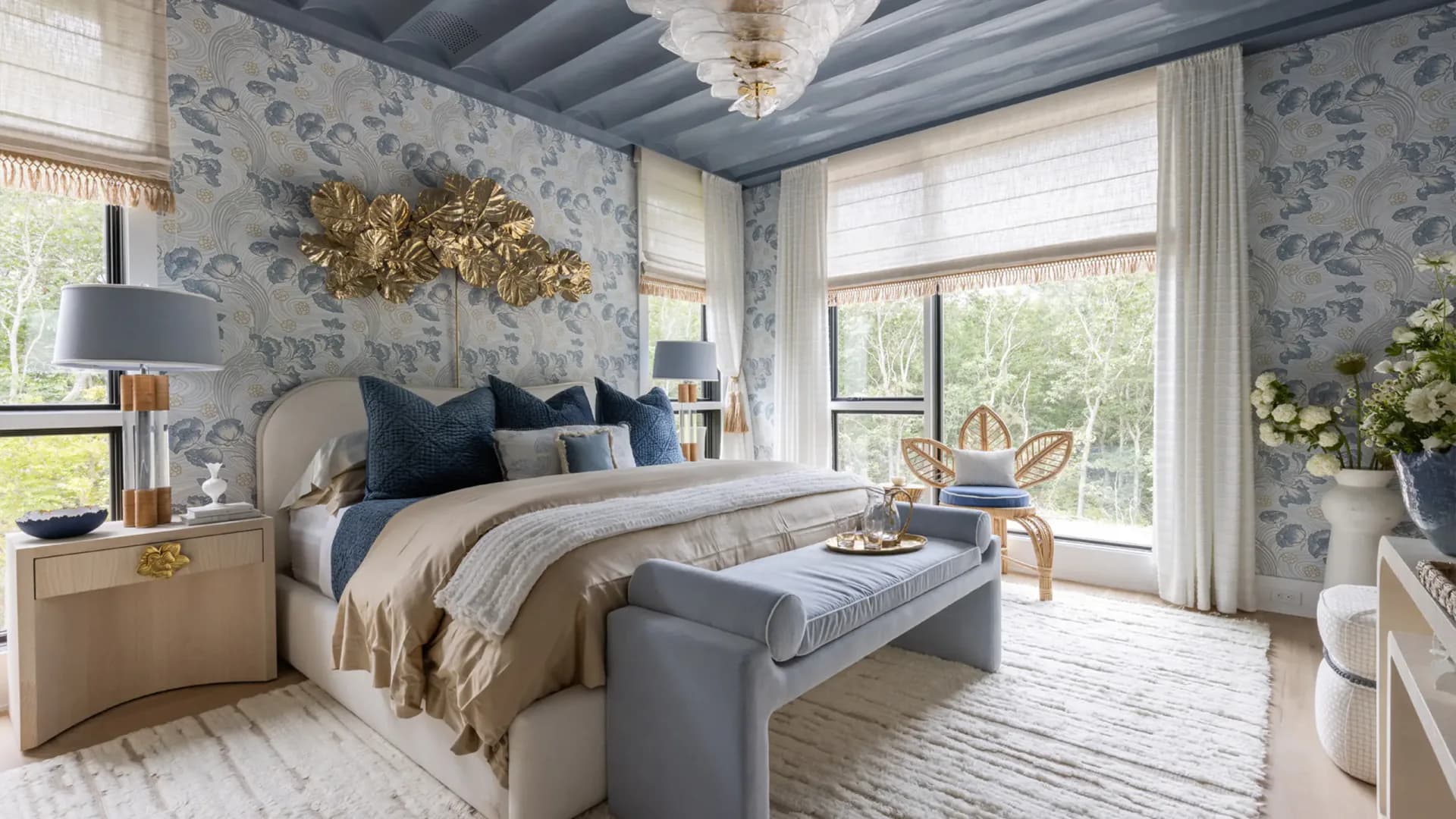 Luxurious primary bedroom with blue floral wallpaper, gray upholstered bed, layered neutral bedding, and French doors opening to gardens by Caroline Zogheb.