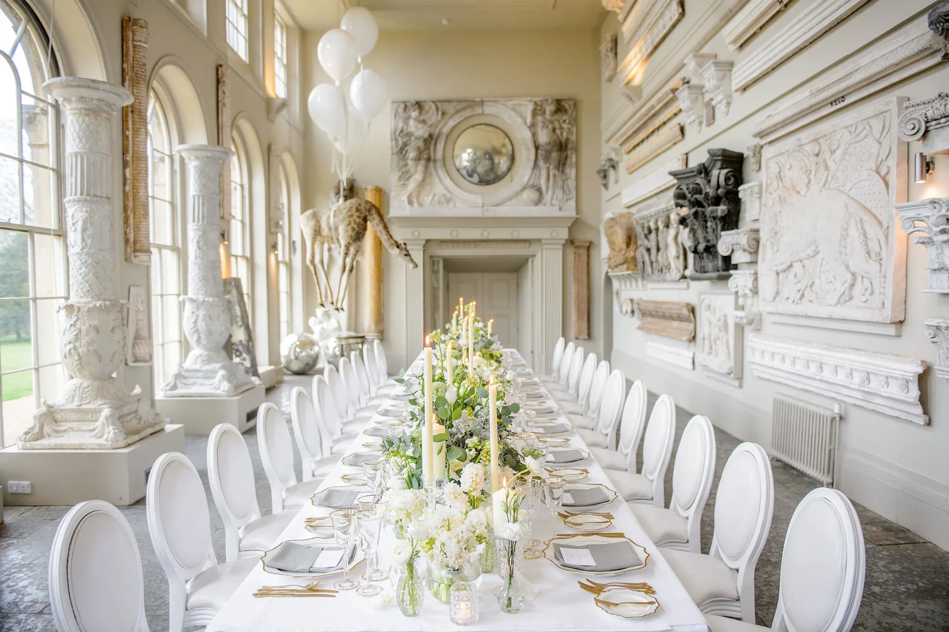 Elegant neoclassical dining room with white sculptural chairs, long table adorned with white floral arrangements, marble fireplace, arched windows, and symmetrical classical details, designed by Cassandre Bonhomme.