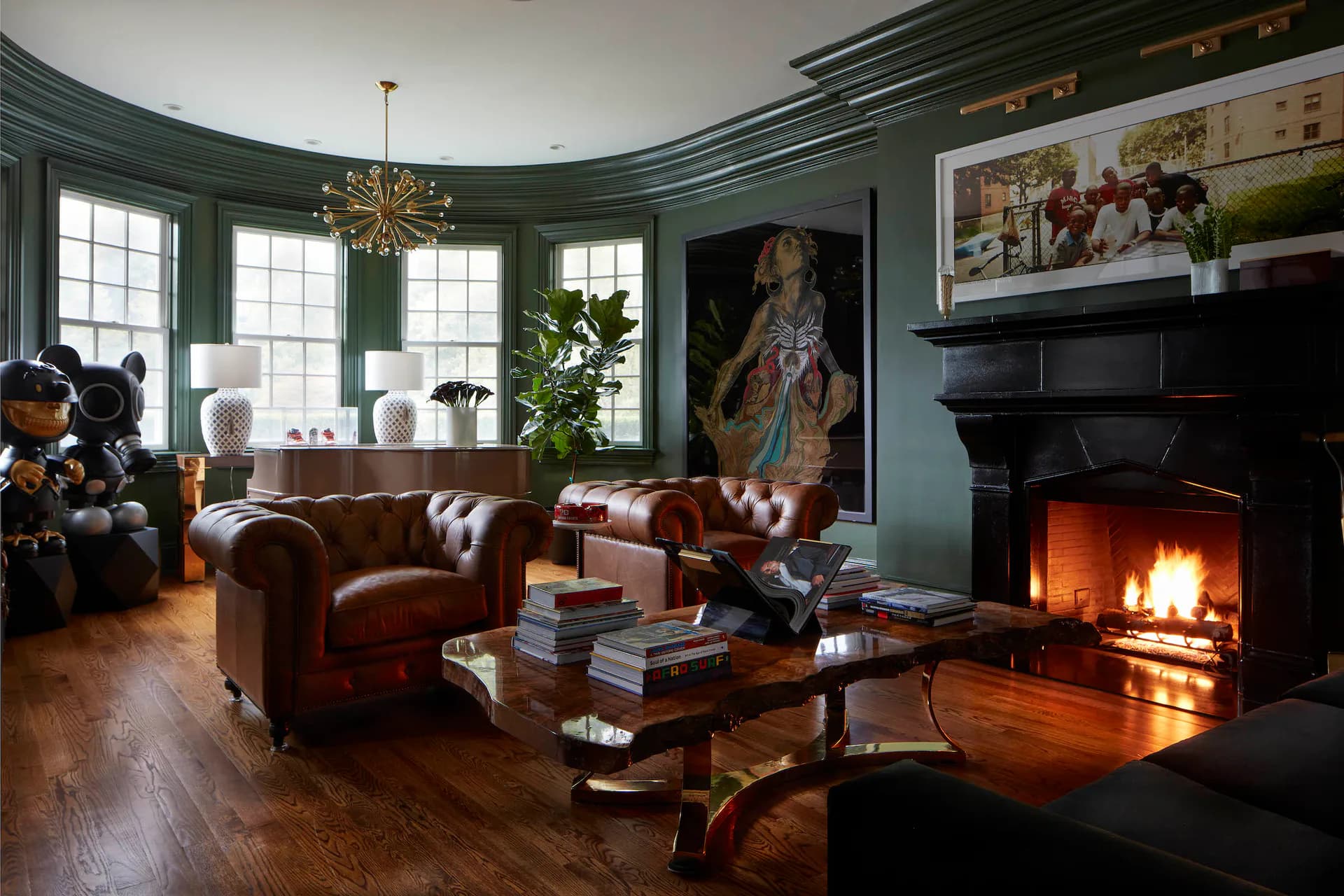 Elegant living room with sage green walls, burgundy leather Chesterfield sofa, fireplace with active fire, bay windows, brass chandelier, and warm wood flooring, designed by Cassandre Bonhomme.