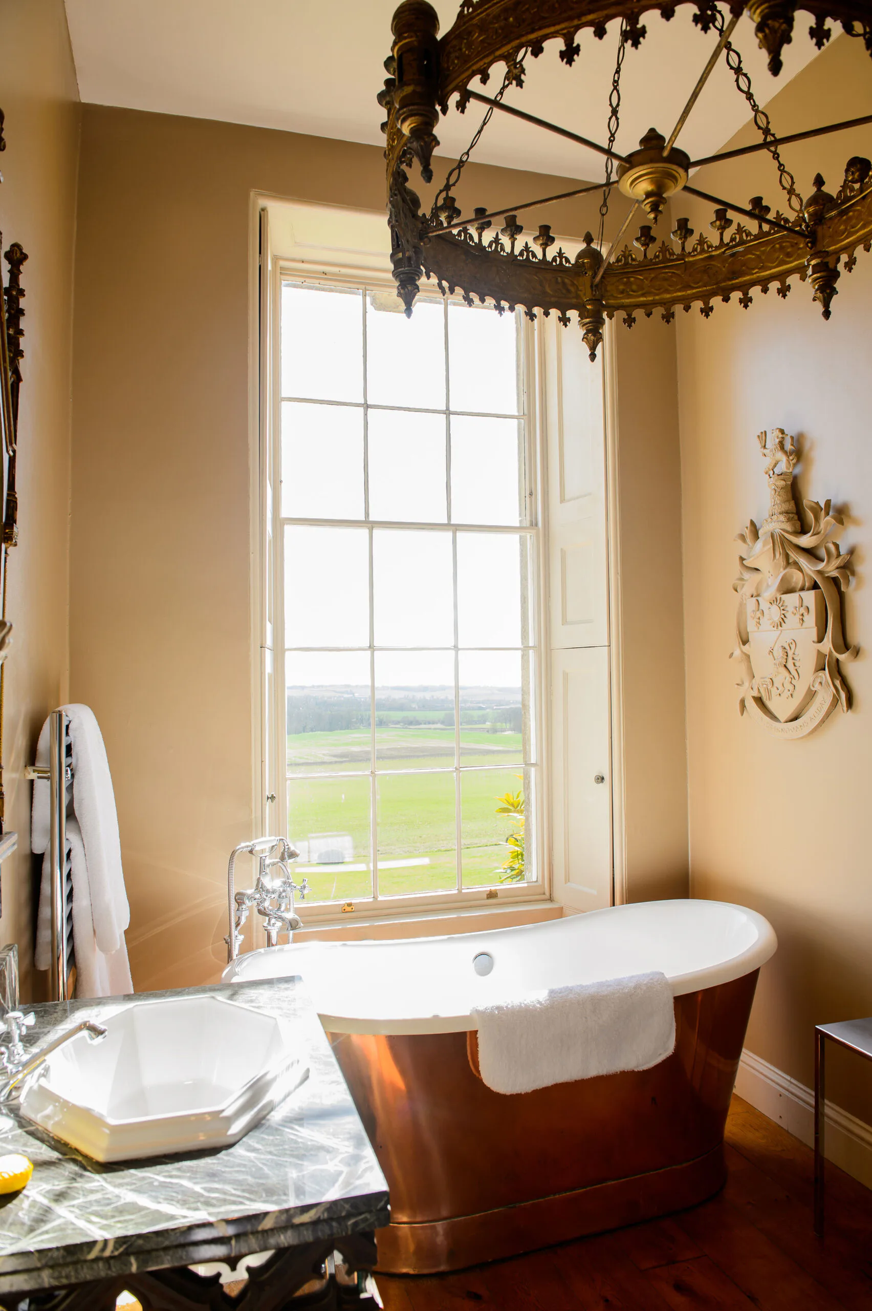 Luxurious bathroom with cream walls, ornate brass chandelier, freestanding copper tub, marble countertop, and tall sash window overlooking countryside by Cassandre Bonhomme.